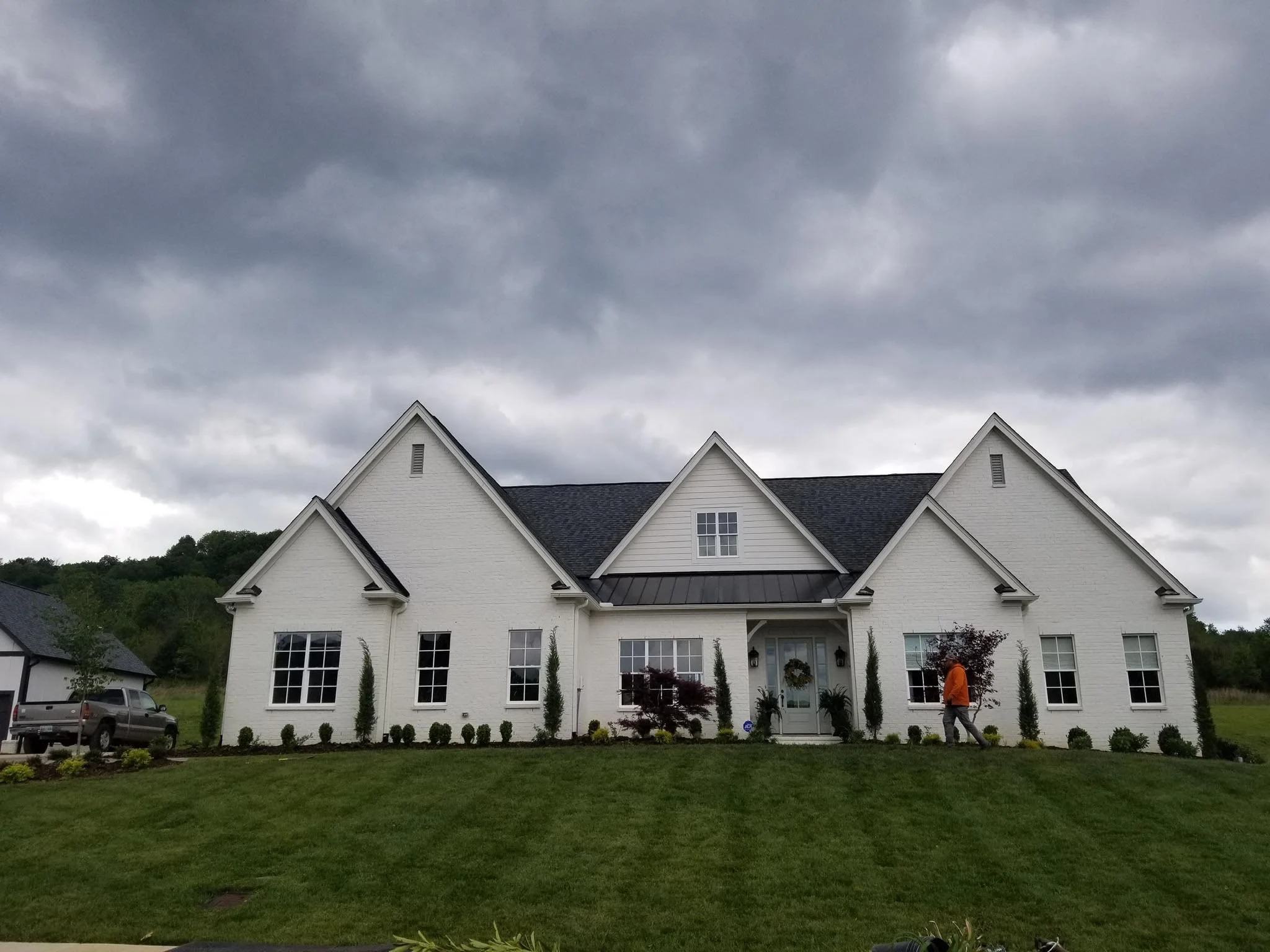 Front view of a large white house with multiple gables and black roofing, surrounded by green lawn and small trees, under a cloudy sky.