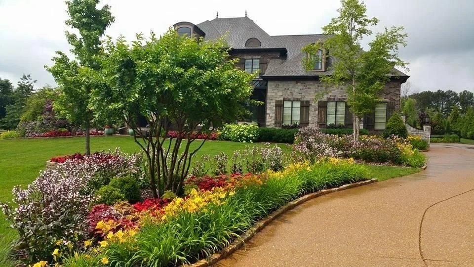 A large house with a dark gray roof is surrounded by a well-manicured yard and colorful flower beds, with a curved driveway leading to it.