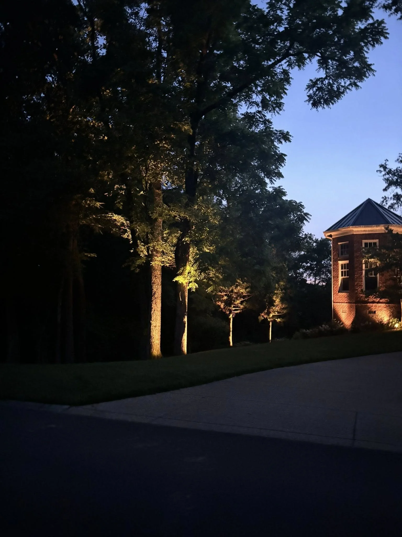 A nighttime view of a residential area with trees illuminated by outdoor lights. A brick house with a turret is visible on the right. The sky is clear with a hint of blue.