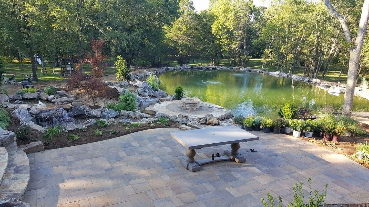 A landscaped backyard with a pool, surrounding trees, a stone fire pit, a paved patio with a table, and potted plants.