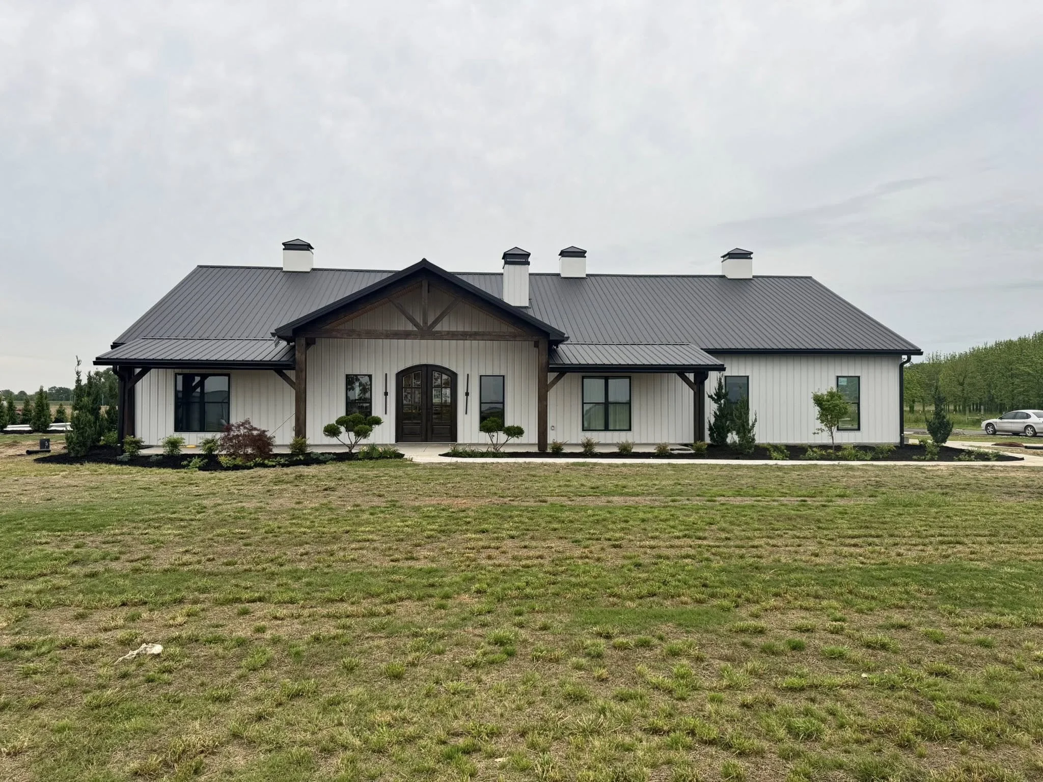 A large modern farmhouse with white exterior walls, black framed windows, and a metal roof surrounded by a landscaped lawn with small trees and shrubs.