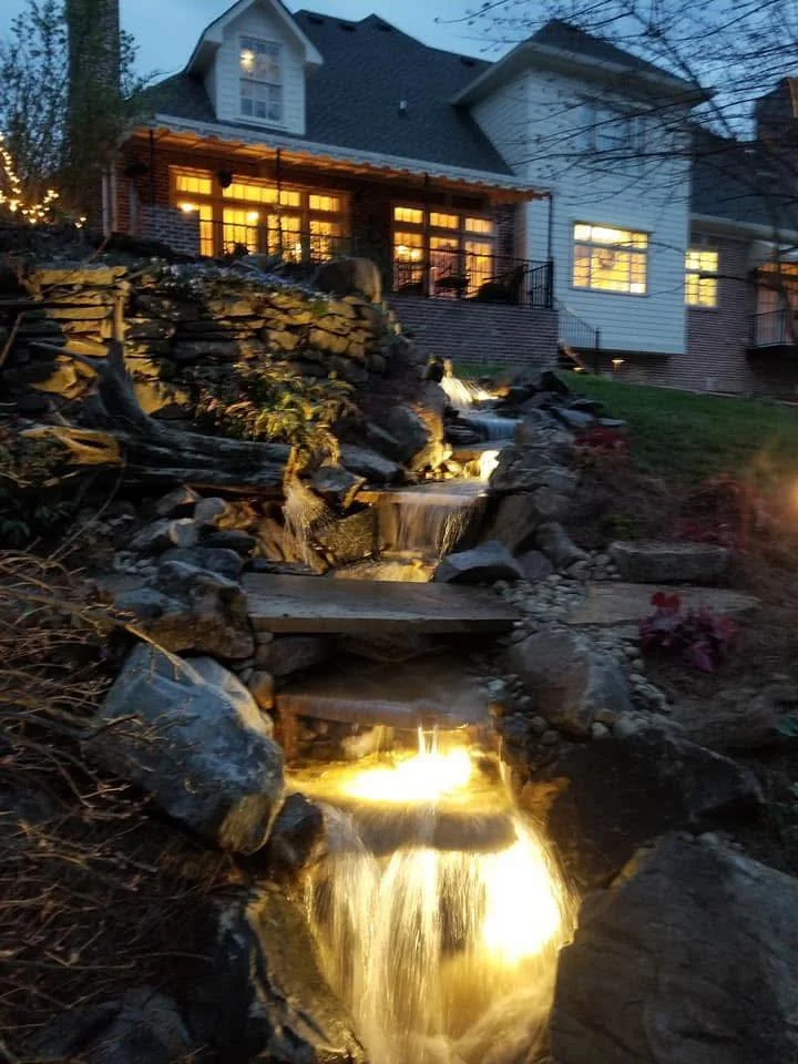 A multi-story house at dusk with lit windows, a balcony, and a garden with a tiered waterfall and stream illuminated with outdoor lighting.