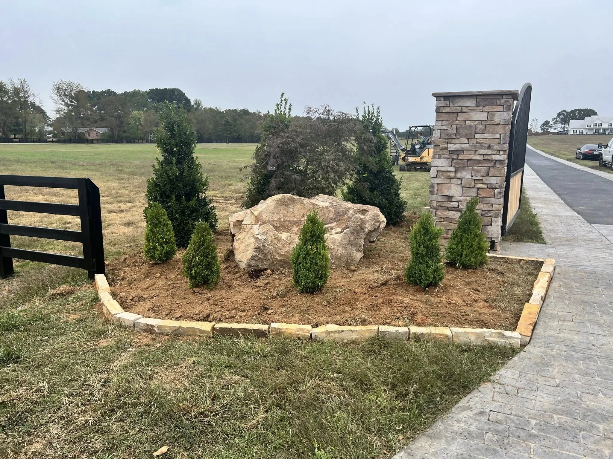 A small landscaped garden bed with shrubs, a large rock, and a stone wall on the right side, bordered by a low stone edge and adjacent to a paved walkway.