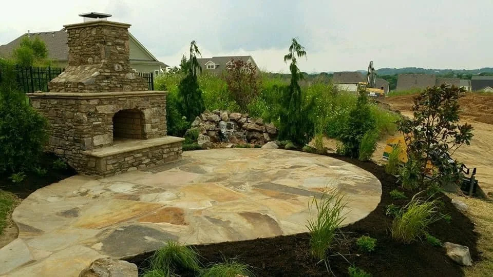 Backyard with a stone outdoor fireplace, a large stone patio, various plants, and construction equipment in the background.