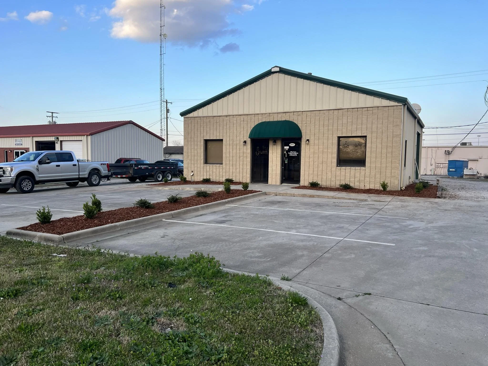 A beige commercial building with a green roof and awning, two windows, and a glass door, surrounded by a parking lot with a few parked trucks and small landscaped shrubs.