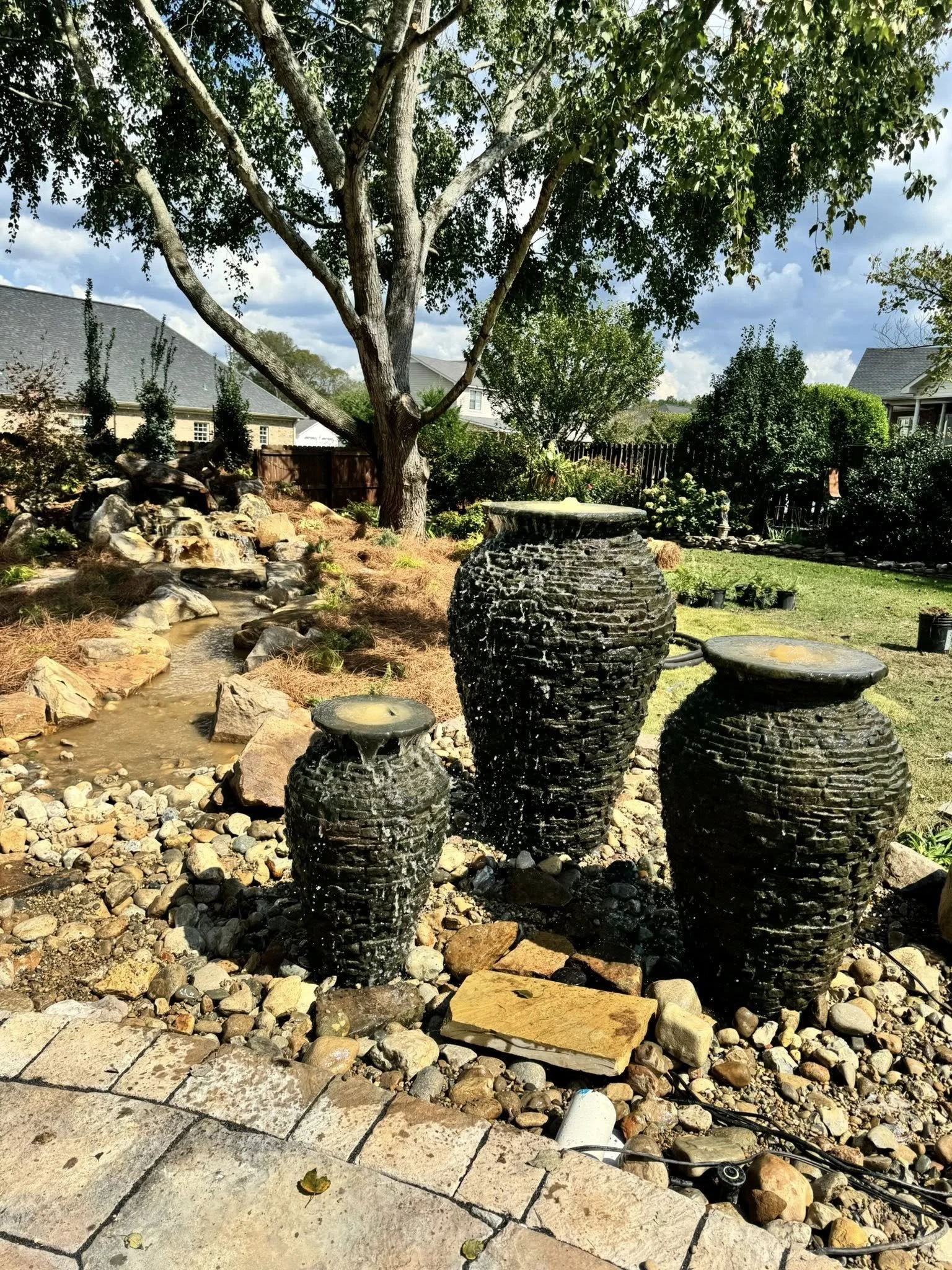 Three black stone outdoor water fountains with water flowing, set among rocks in a landscaped backyard with a large tree, a small creek, and a wooden fence.