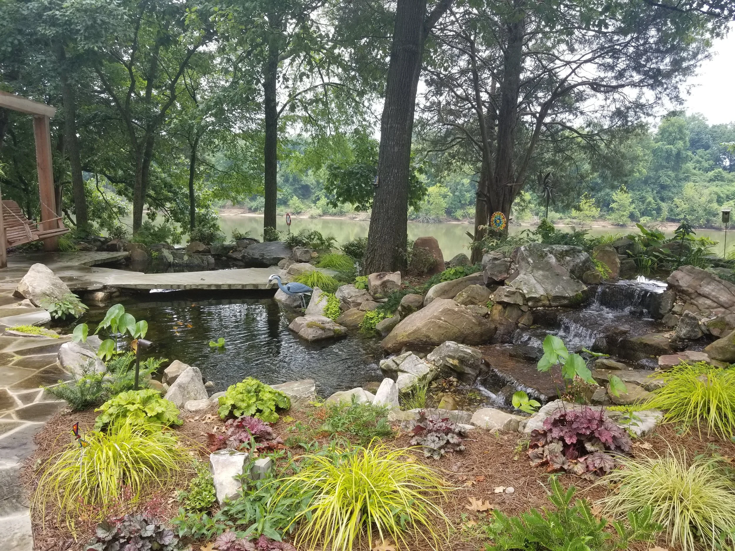 A serene backyard garden featuring a small pond with lily pads, a stone pathway, lush green plants, rocks, and trees overlooking a river in the background.
