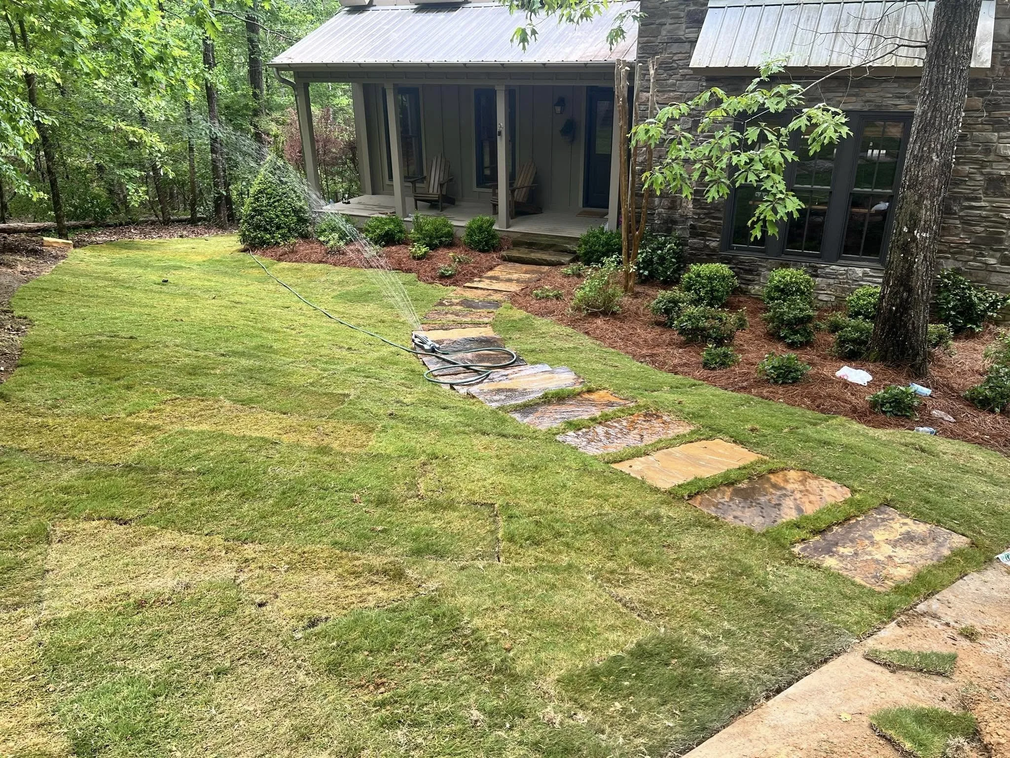 Garden lawn being watered with a sprinkler, stone pathway leading to a porch with chairs, surrounded by shrubs and trees, outside a house with stone and siding exterior.