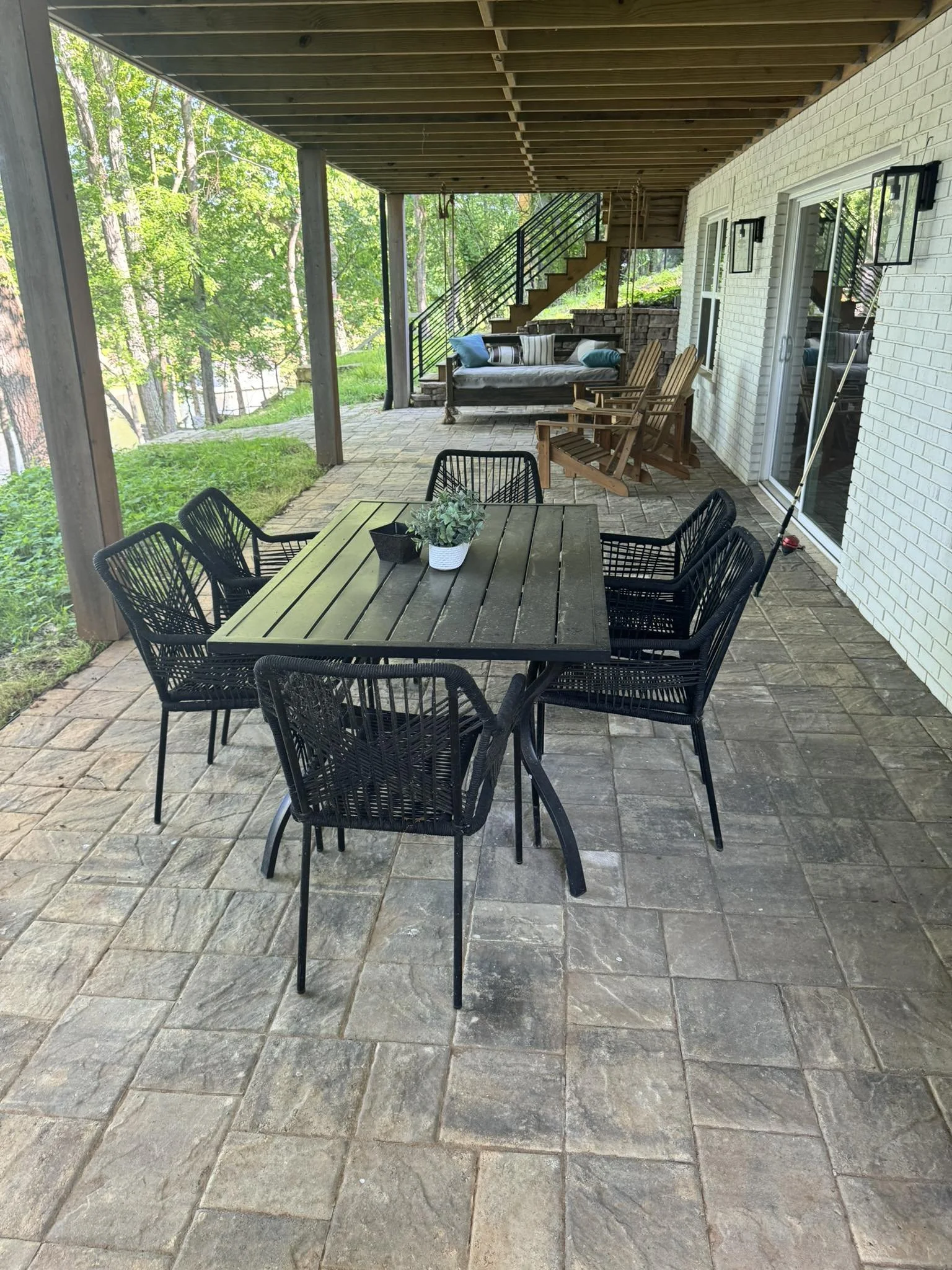 Outdoor patio with a wooden dining table and six black chairs, surrounded by a wooded area, with a white brick house wall and outdoor furniture including rocking chairs and a sofa in the background.