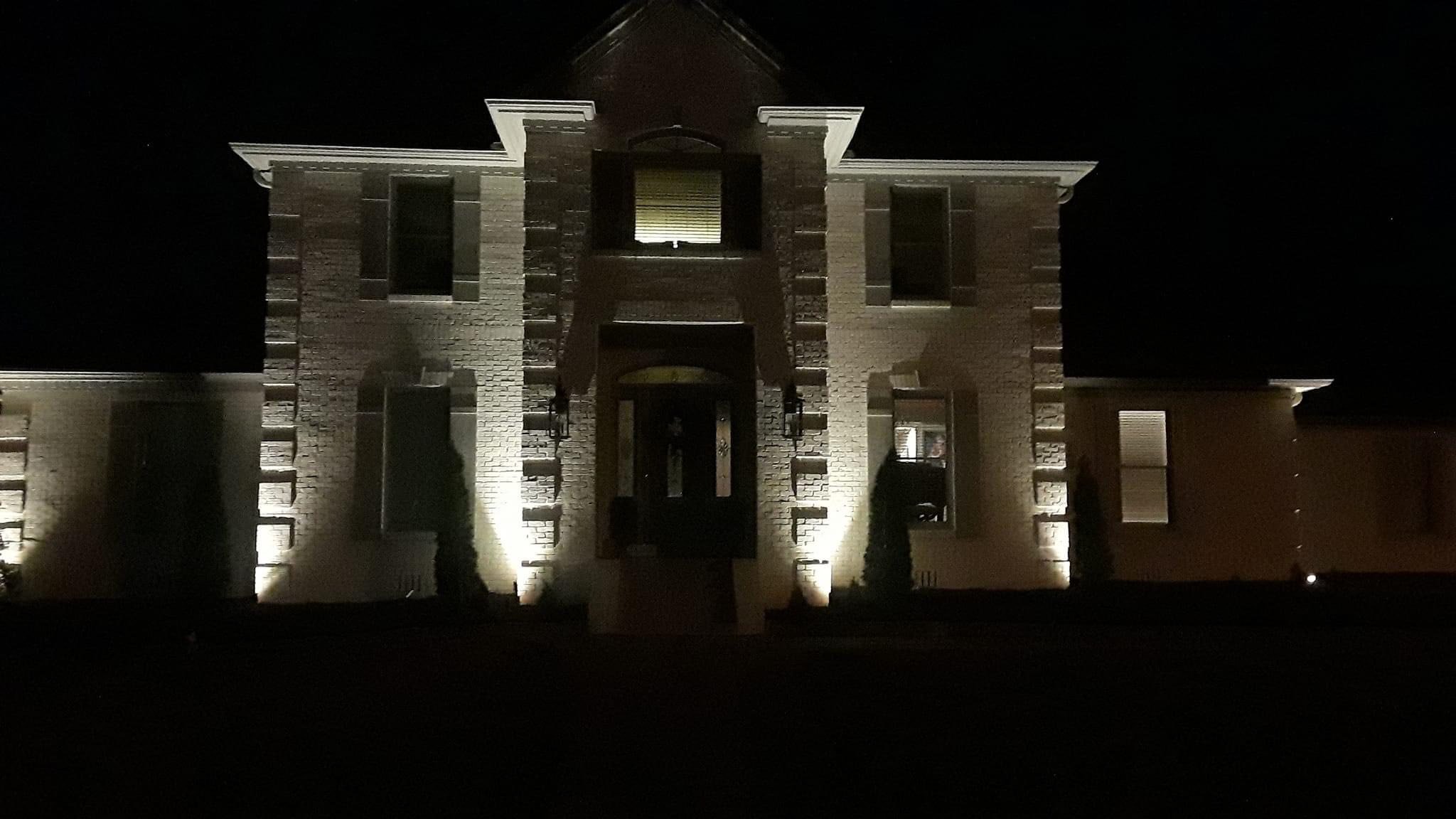 A two-story brick house illuminated by exterior lights at night, featuring black shutters, a front door, and tall, narrow windows.