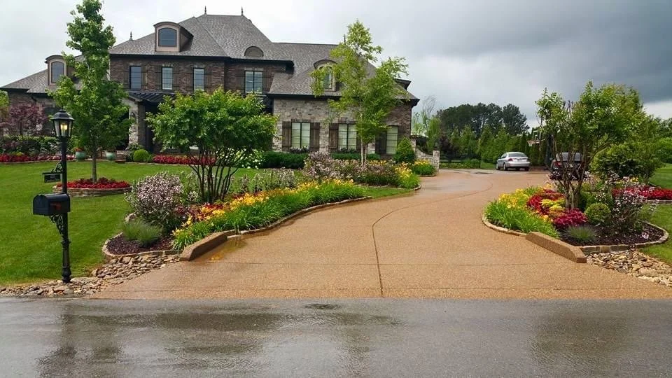 A residential house with a well-maintained front yard, lush green lawn, colorful flower beds, and a curved driveway with a car parked in the background. The sky is overcast.