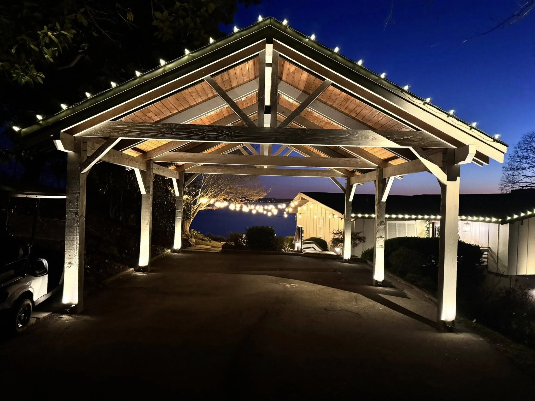 Wooden carport illuminated with string lights at night with a small white building and trees in the background.