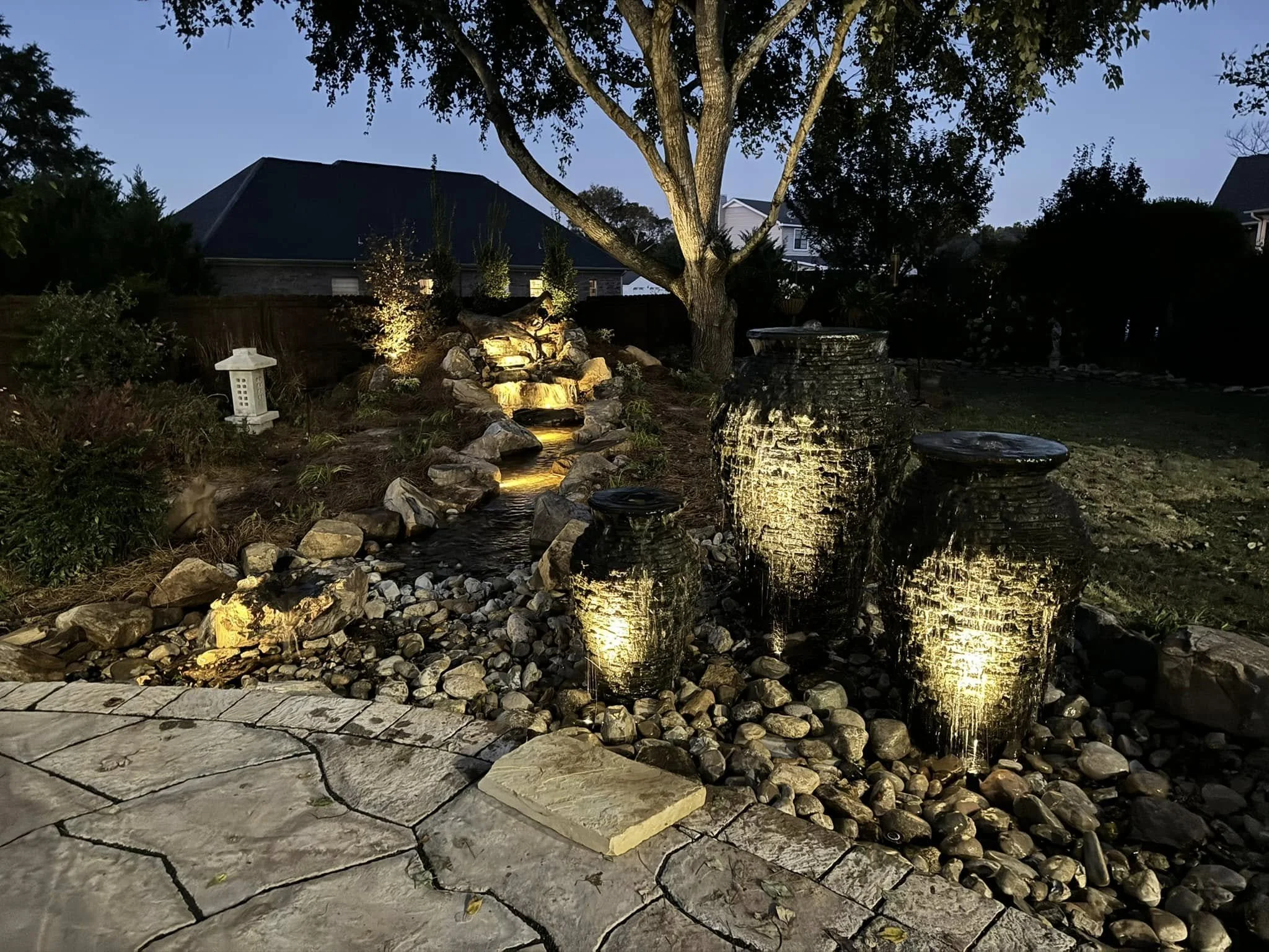 Backyard garden with a small waterfall feature illuminated at night, surrounded by rocks, large decorative planters, and outdoor lighting, with trees and a house in the background.