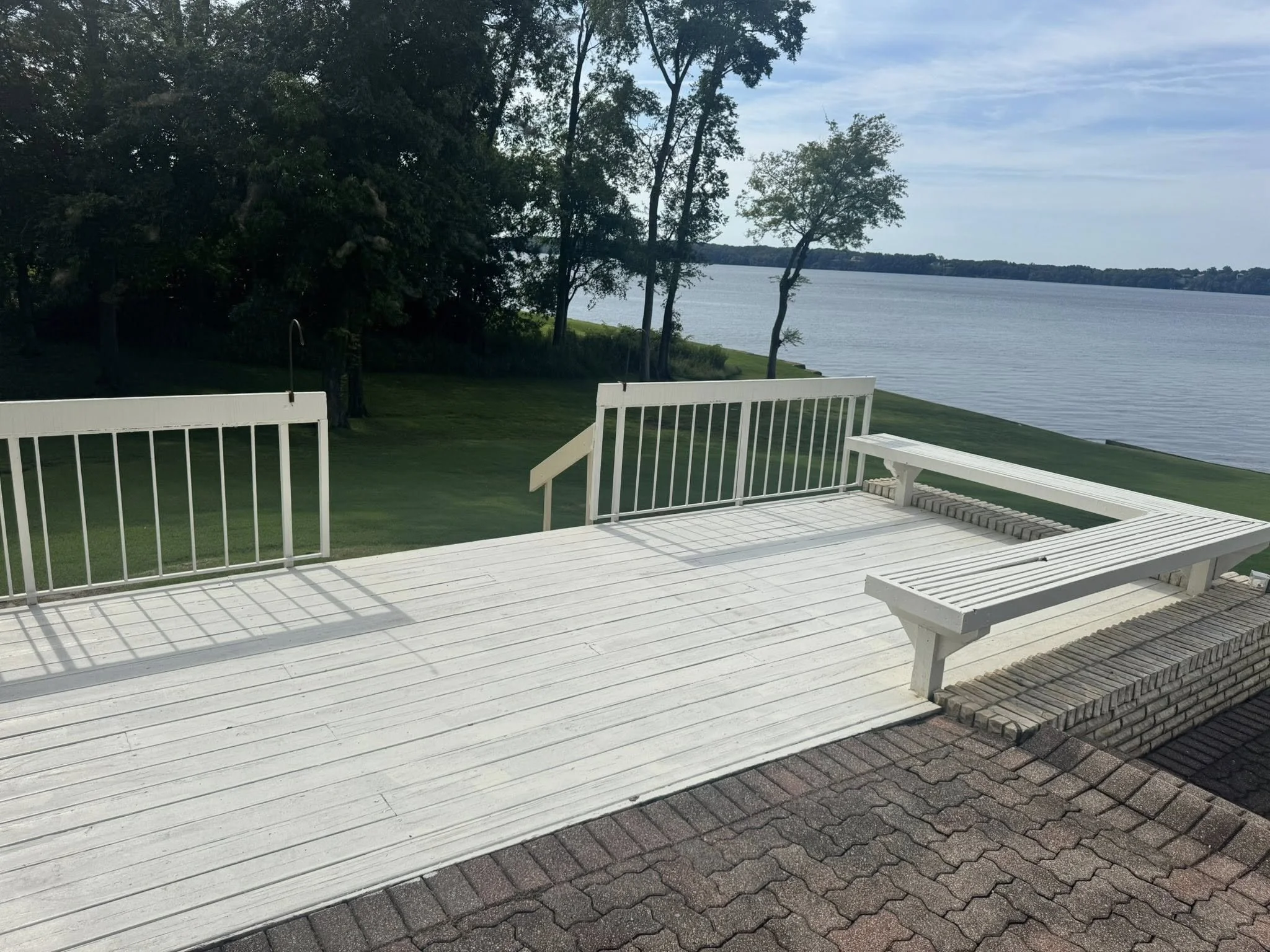 White wooden deck with built-in bench overlooking a lake, with trees and grassy shoreline in the background.