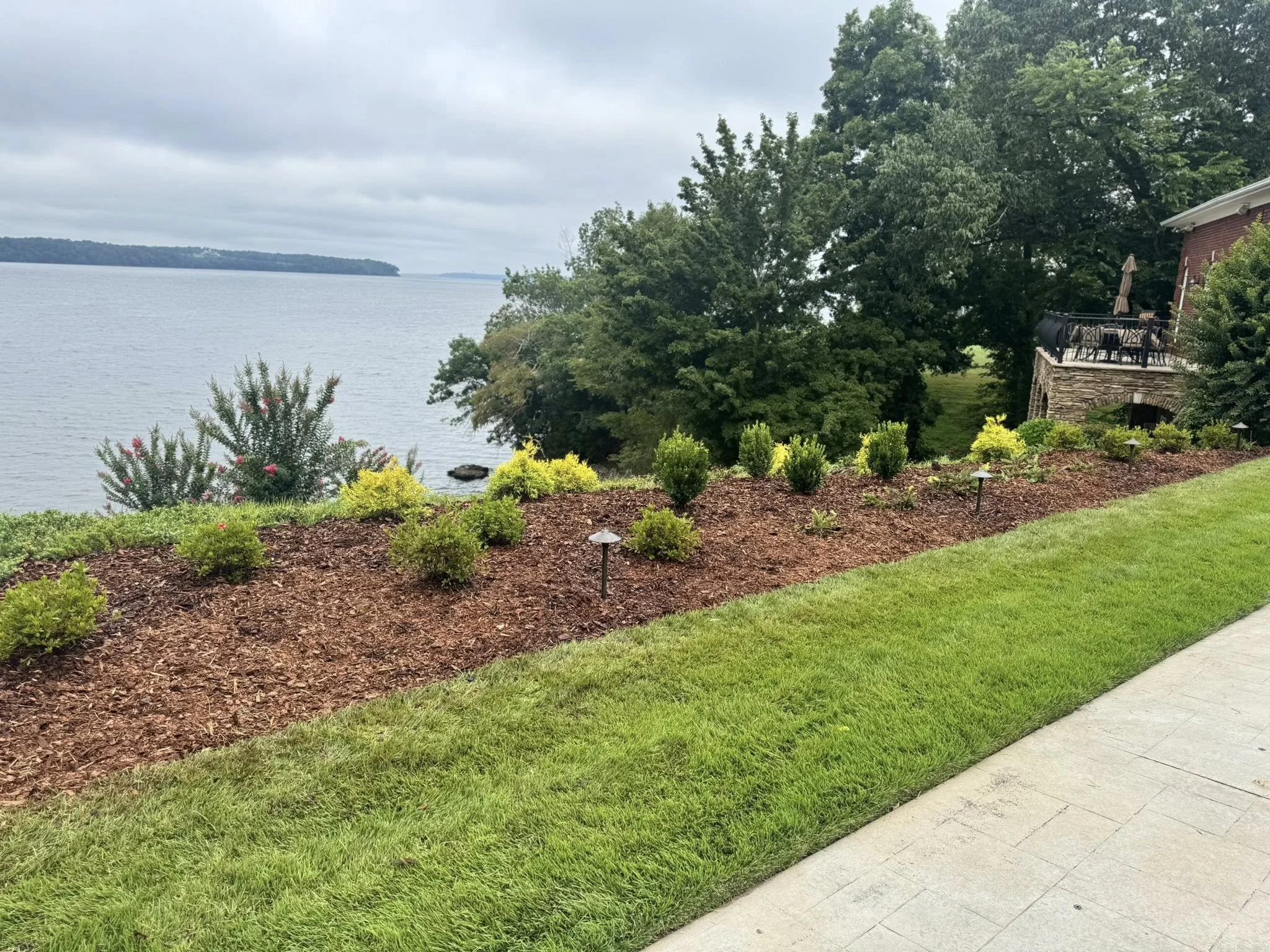 A landscaped garden with small green shrubs and pink flowering plants along a lakeside, with a stone patio and a house with a balcony in the background, surrounded by lush trees on a cloudy day.