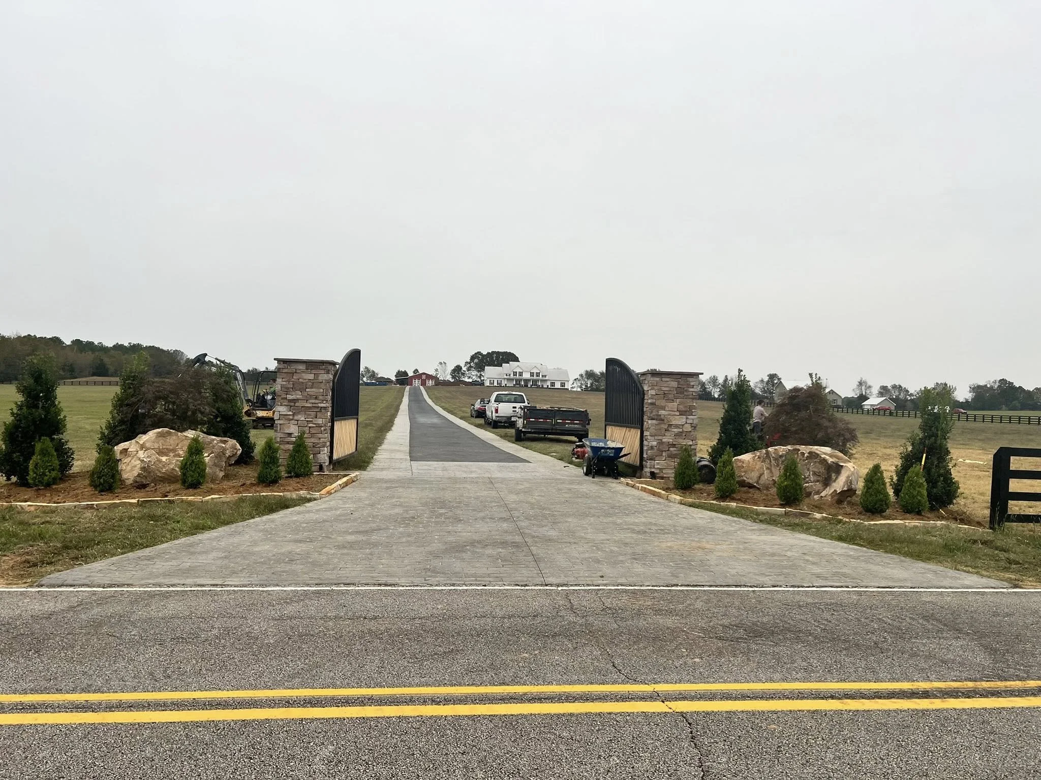 Driveway entrance with stone and black gate, landscaped with small trees and rocks, leading to a rural property with open fields and distant buildings.