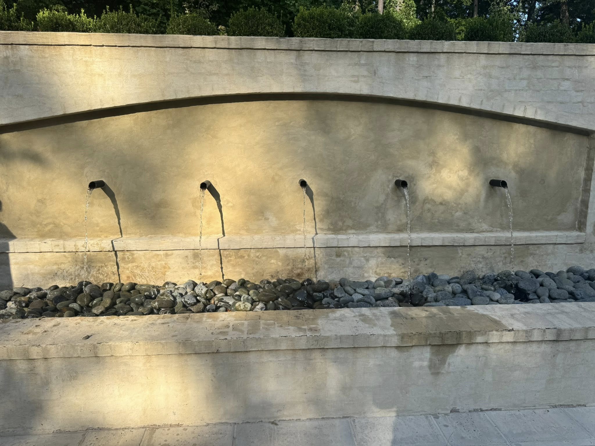 A decorative outdoor water fountain with five streams of water flowing from pipes into a bed of smooth stones.