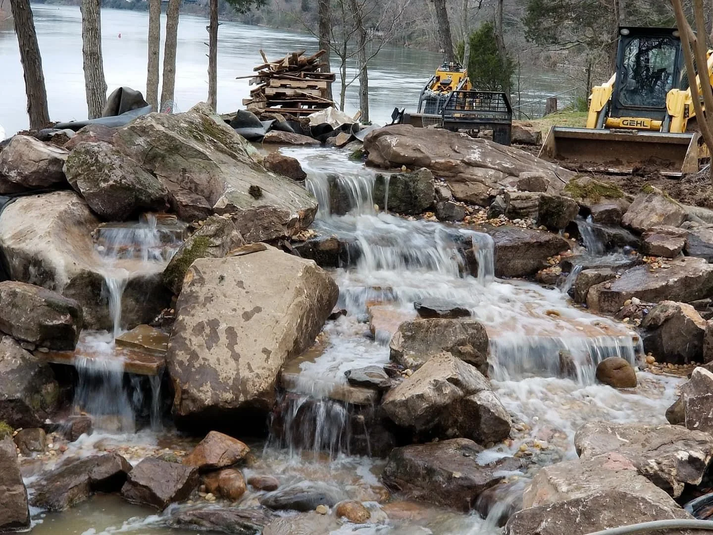 Construction site on riverbank with rocks and small waterfall, construction equipment including bulldozer and machinery near water, surrounded by trees.