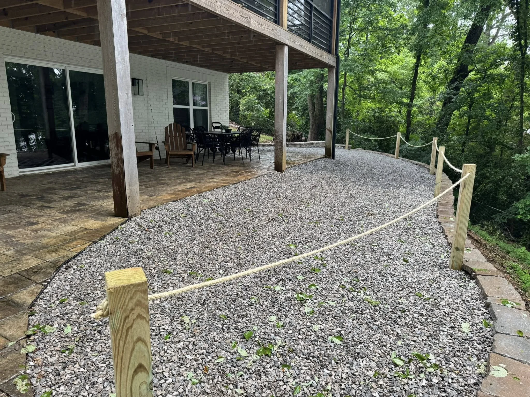 Covered outdoor patio with wooden posts, a gravel walkway, and a railing, adjacent to a house with sliding glass doors, surrounded by green trees.