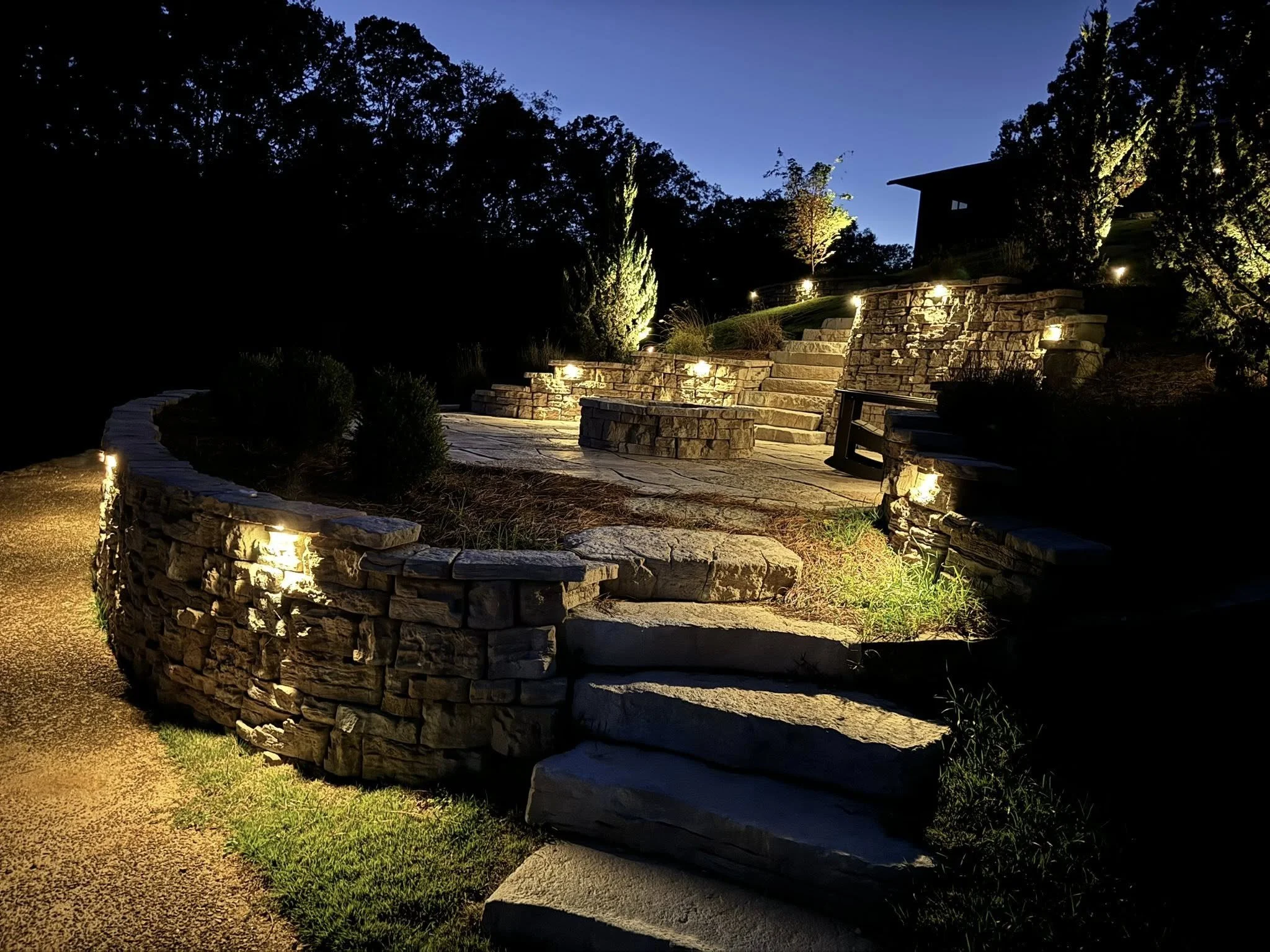 Night view of a landscaped backyard with stone steps and retaining walls illuminated by pathway lights, trees, and a house in the background.