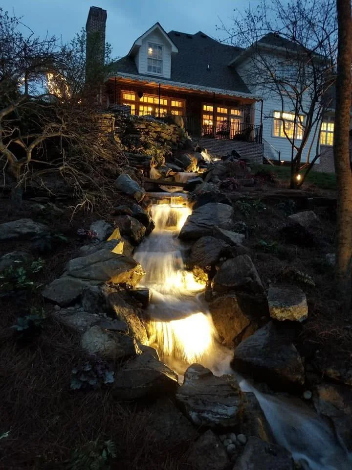 A house at dusk with a waterfall feature flowing down rocks in the foreground, illuminated by outdoor lighting, with a porch and trees around.