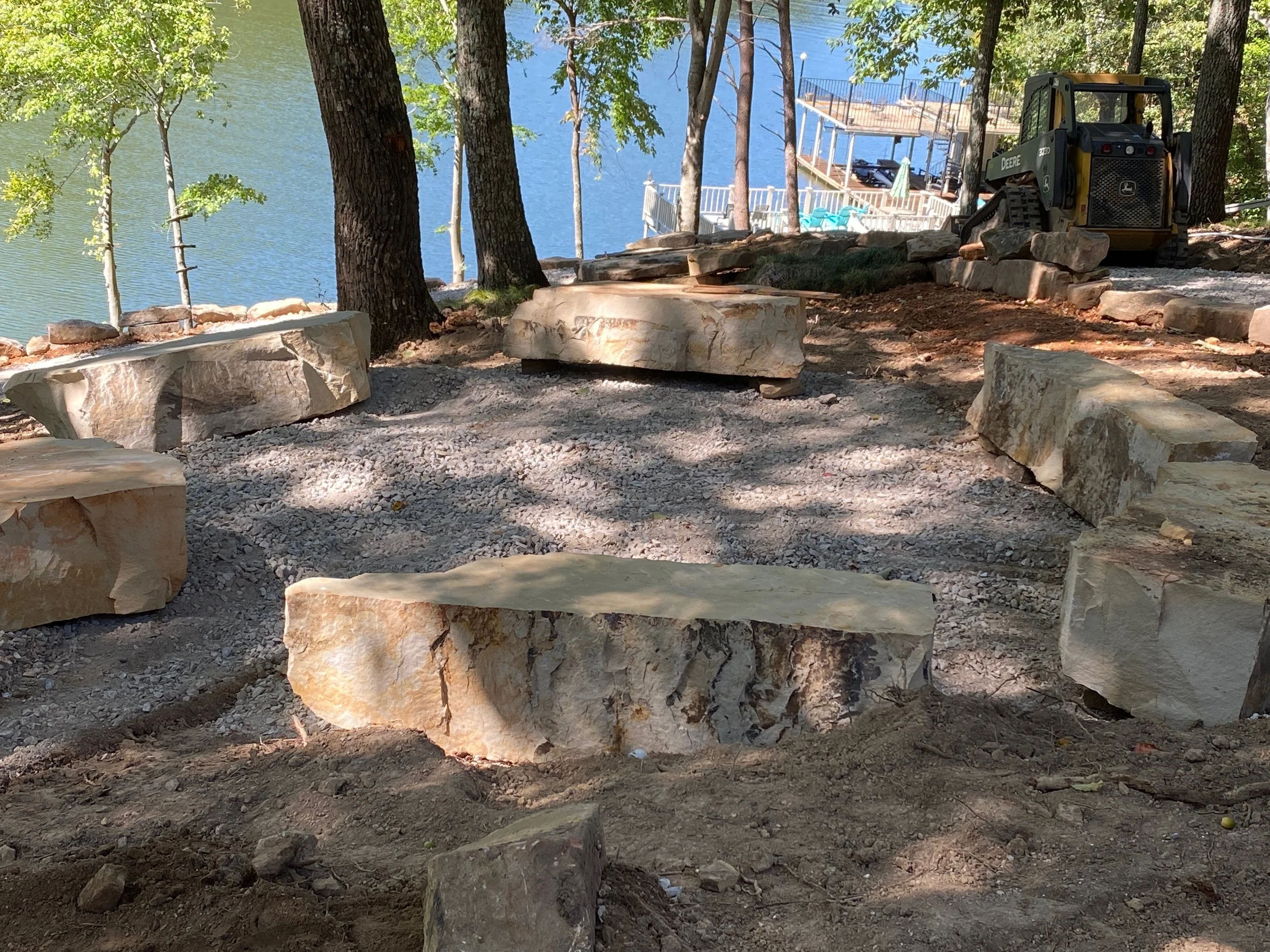 Large stones being arranged for a patio or pathway near a lake with trees and a boat dock in the background, and construction equipment present.