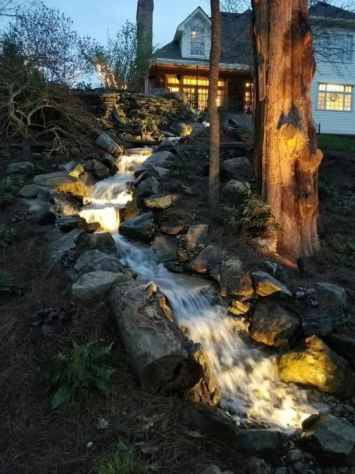A backyard with a small waterfall or stream flowing over rocks, with trees and a house in the background illuminated at dusk.