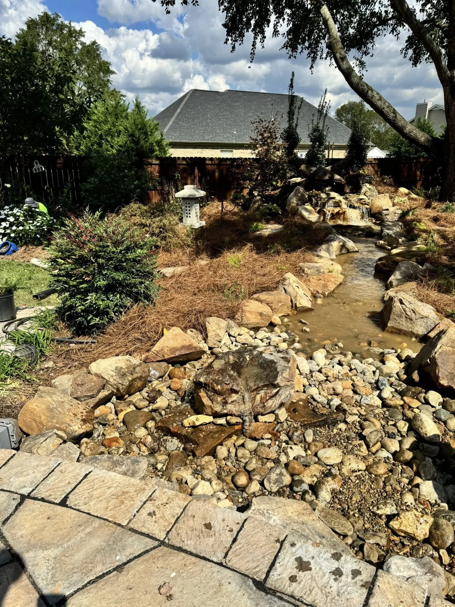 A backyard with a small artificial stream flowing over rocks, surrounded by various plants, flowers, and trees; a stone pathway in the foreground, a lantern, a bird feeder, and a wooden fence visible in the background under a partly cloudy sky.