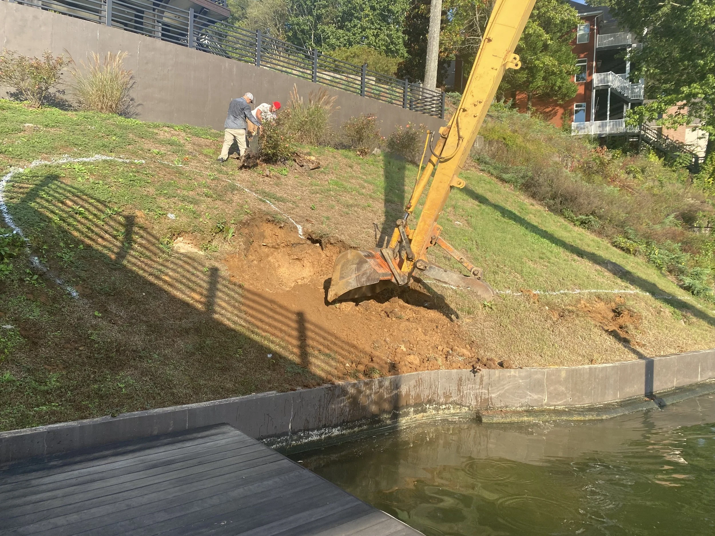 Two workers and an excavator working on hillside near water, with a residential building in the background.