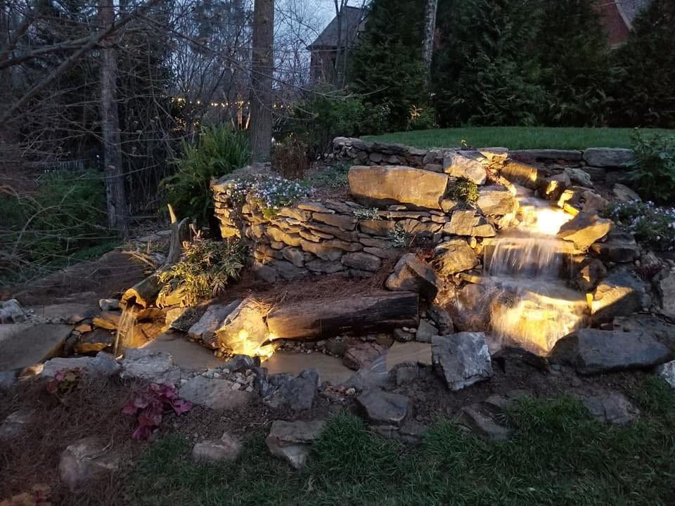 A garden waterfall feature with rocks illuminated by warm lighting, flowing into a small pond surrounded by plants and trees at dusk.