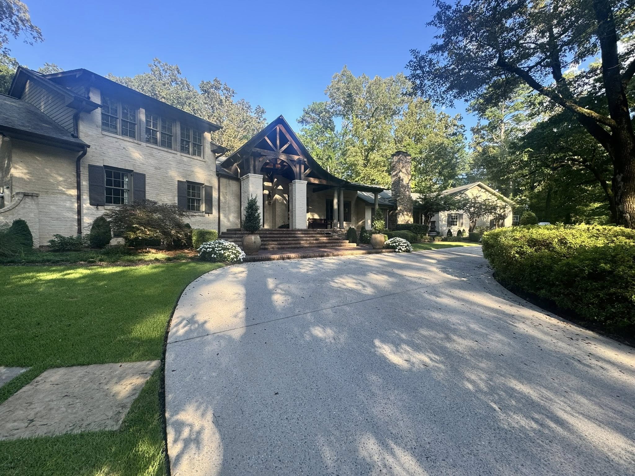 A large, elegant house with brick and stone exterior, surrounded by greenery and trees, with a curved driveway leading to the front entrance.