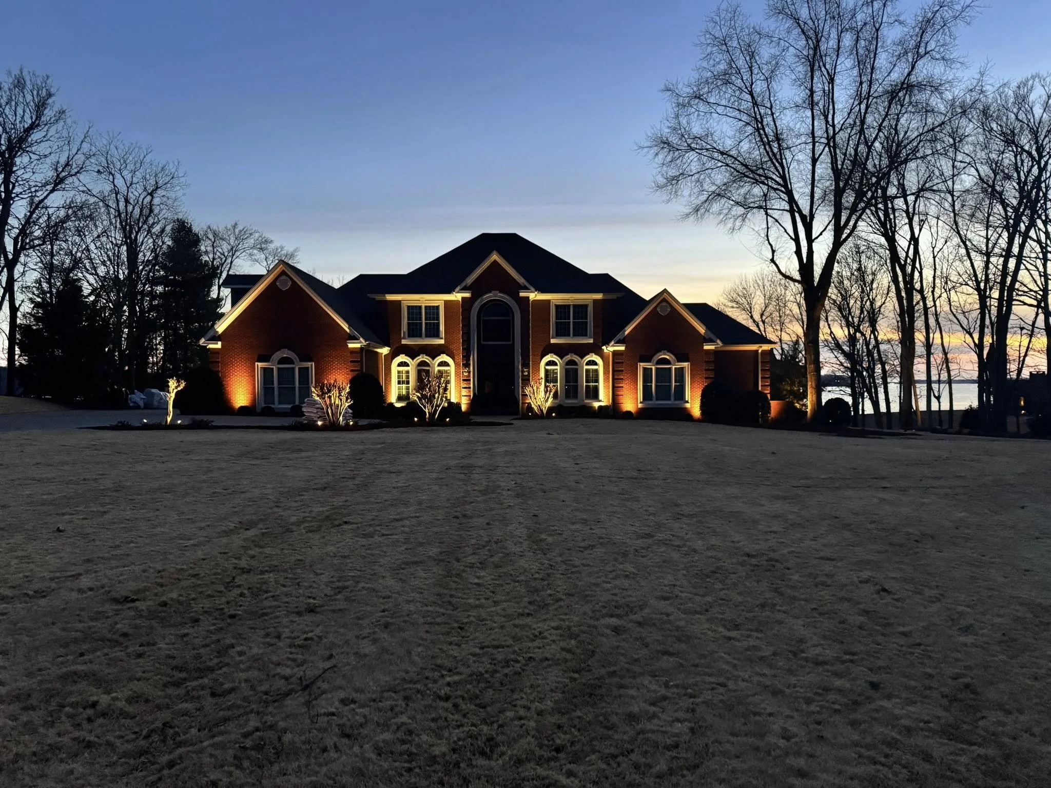 A large brick house illuminated at dusk, with trees and a body of water in the background.