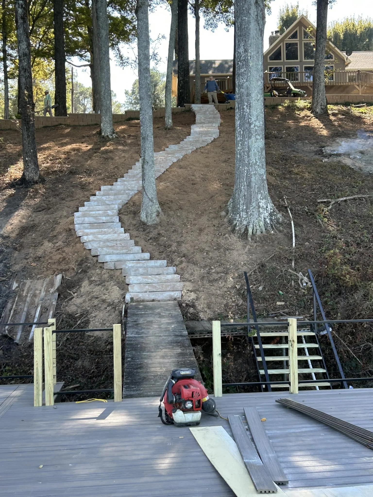 Outdoor construction site with a newly built stone staircase winding up a wooded hill to a house. Construction tools and materials are present on the deck in the foreground.