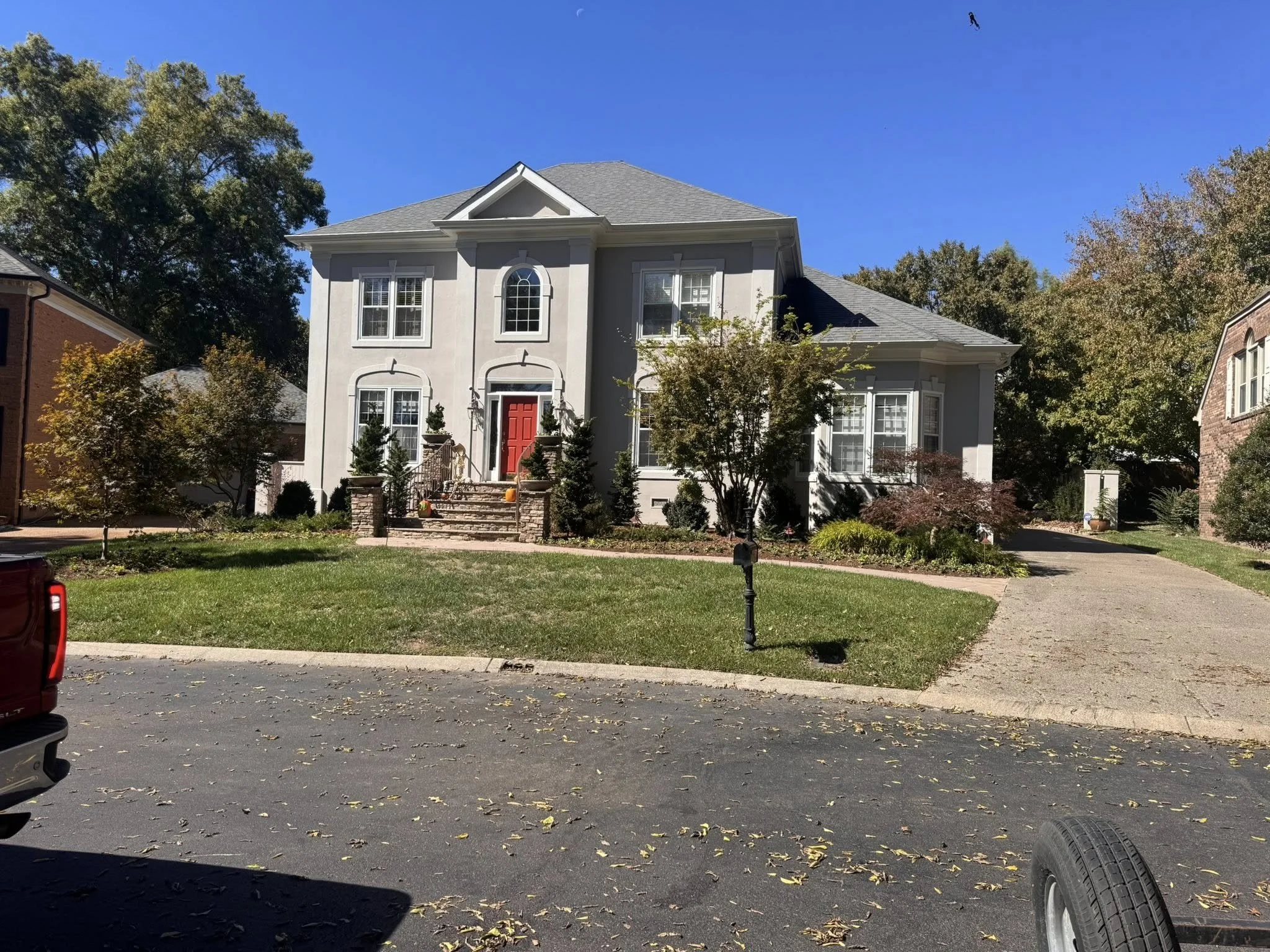 A large two-story house with a gray exterior and a red front door. The house has tall windows with white trims, a front pathway, and steps leading up to the entrance. There are trees and shrubs in the front yard, with a driveway on the right side. Th