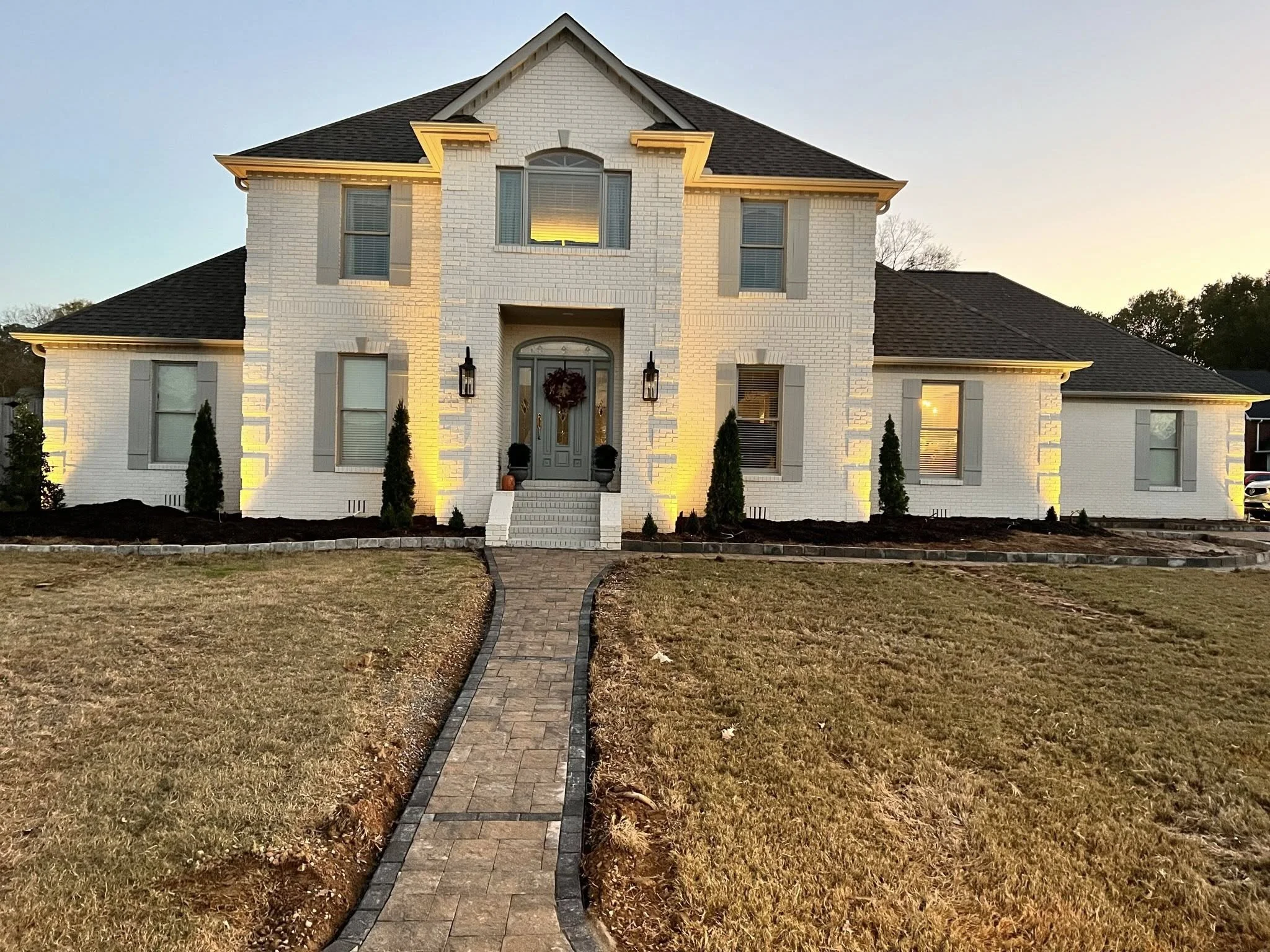 A large two-story white brick house with a front porch, steps, and a curved brick walkway leading to the entrance, illuminated with exterior lights at sunset.