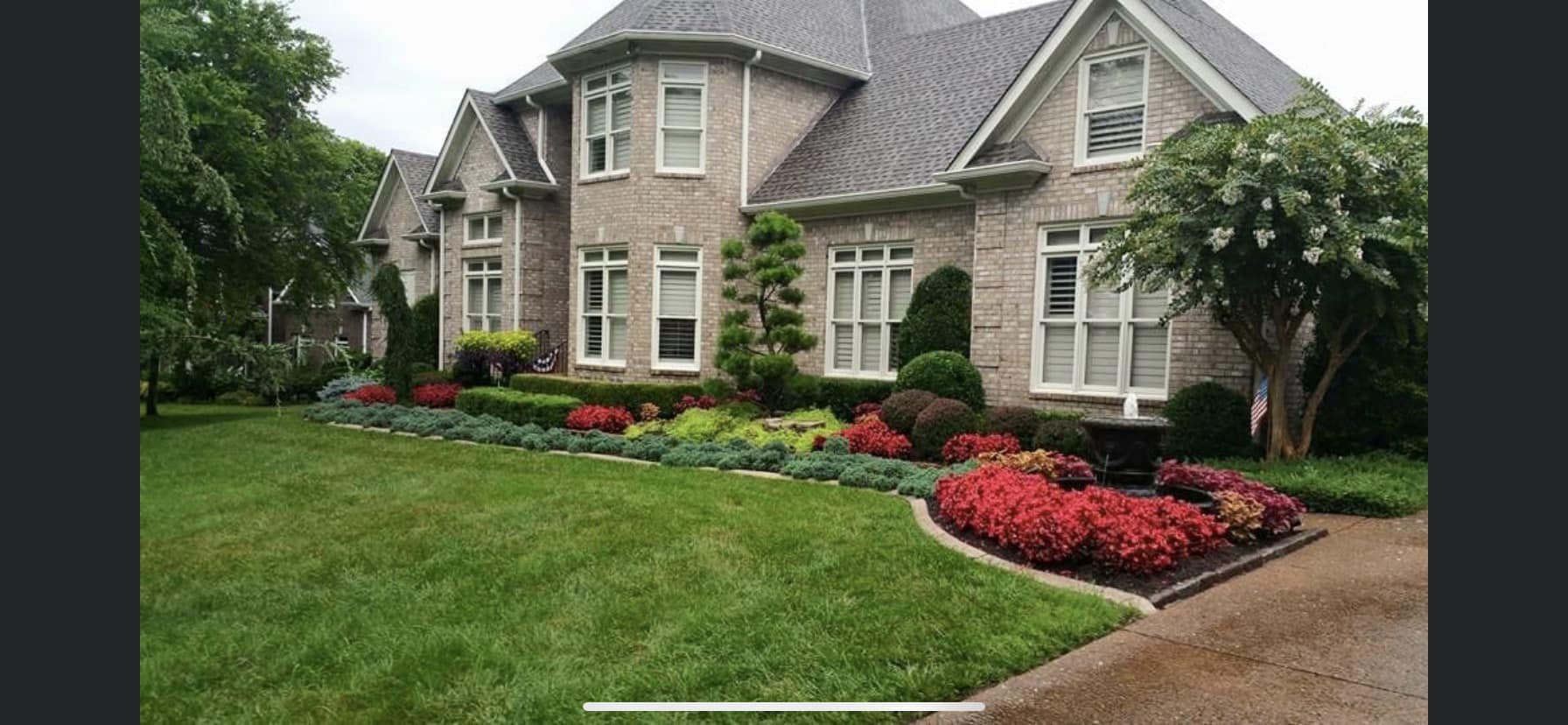 A large brick house with multiple windows and a well-maintained front yard garden featuring a variety of colorful flowers and trimmed bushes, with mature trees on each side.