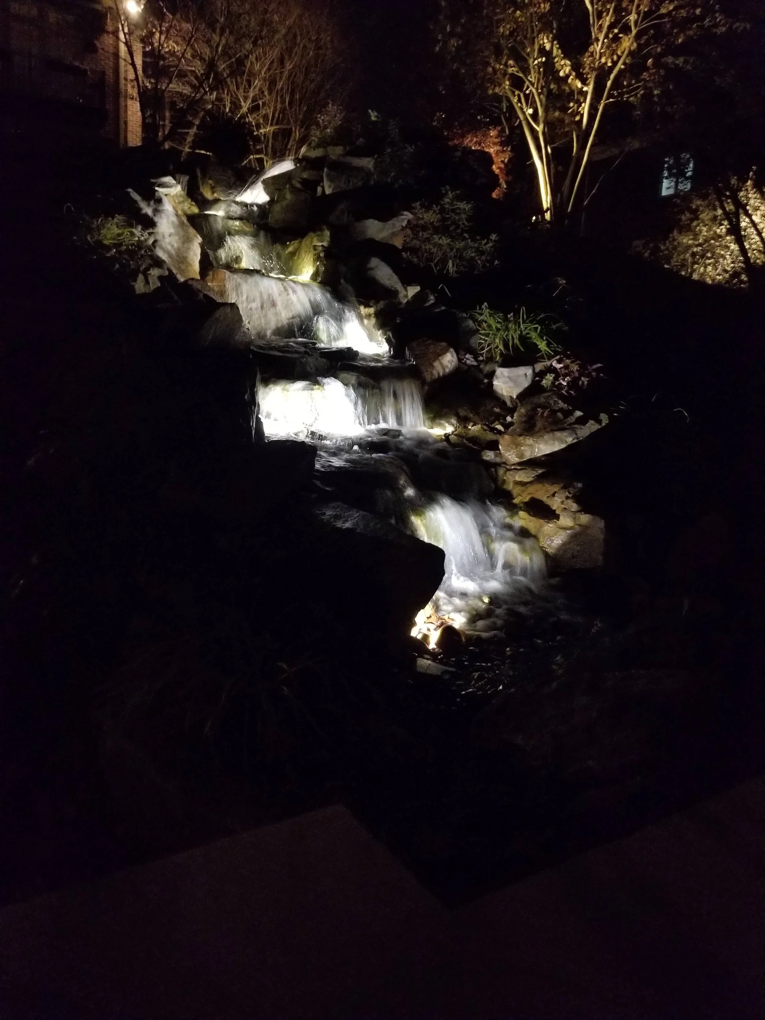 Nighttime view of a small waterfall with rocks and plants, illuminated by outdoor lights, with trees and possibly a building in the background.