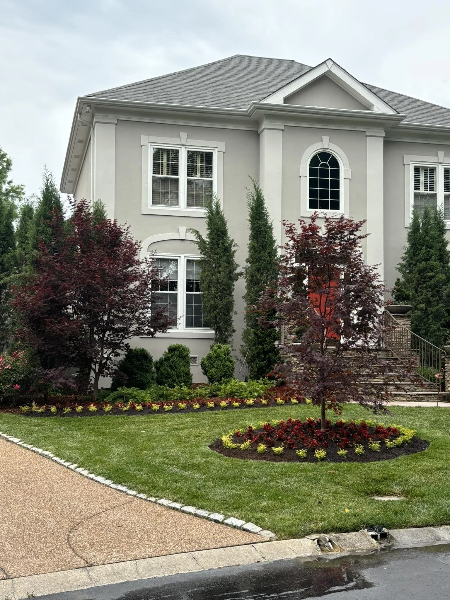 Front view of a large, white, two-story house with multiple windows, a staircase, and a well-maintained garden with trees, shrubs, and flowers.
