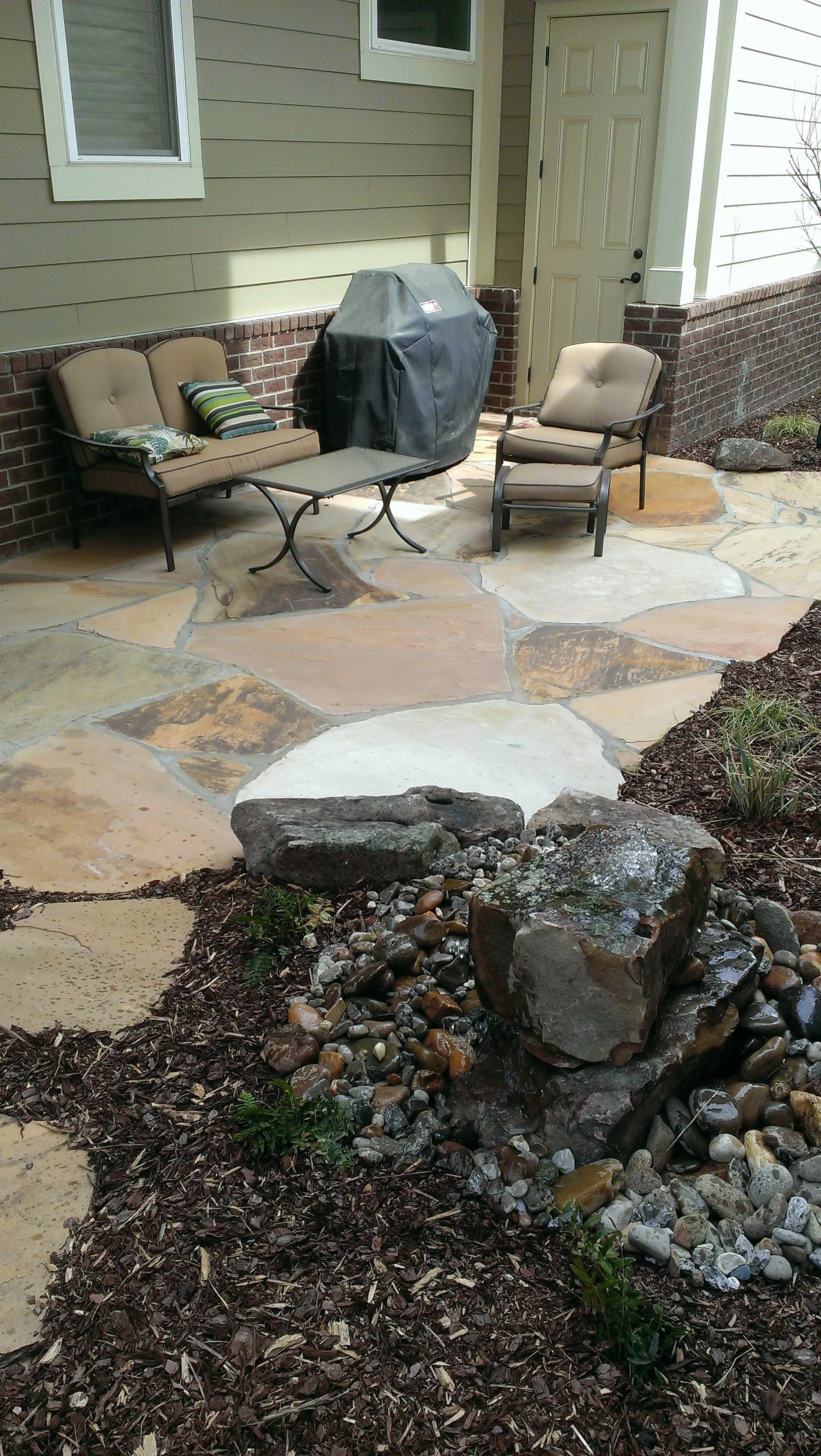 A backyard patio with stone paving, outdoor furniture, a grill covered with a black cover, and a rock garden with large stones and smaller pebbles.