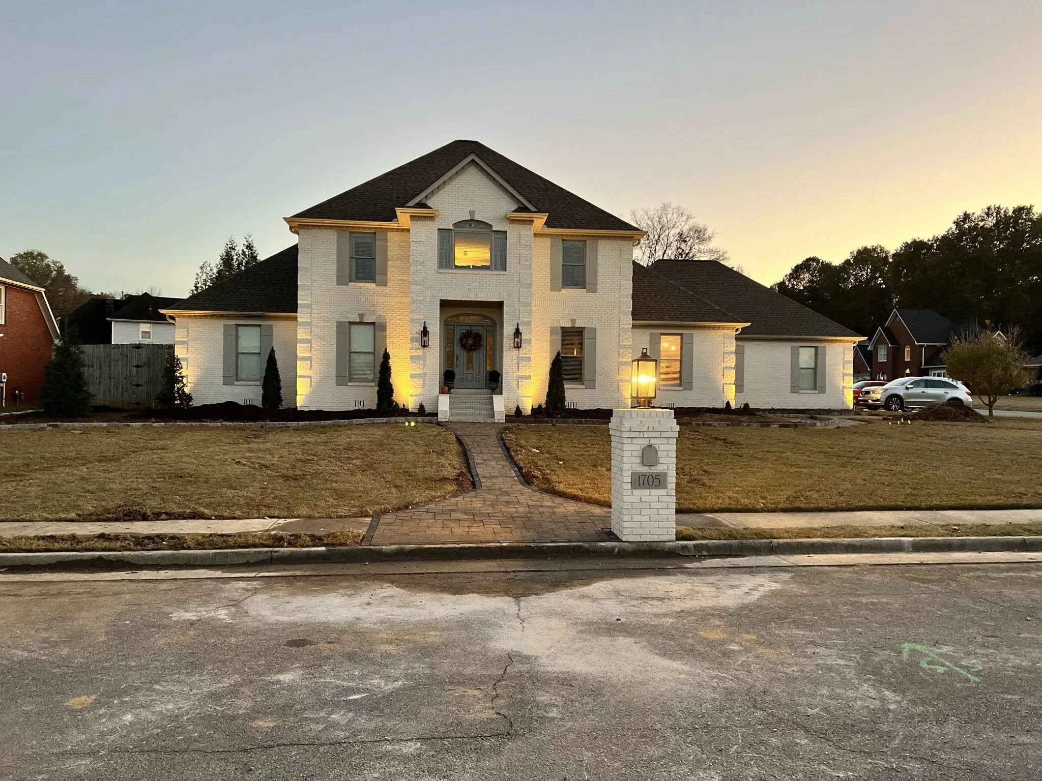 White two-story house with illuminated exterior lights, a small front yard, a brick walkway leading to the front door, and a mailbox with the number 1705, during sunset.