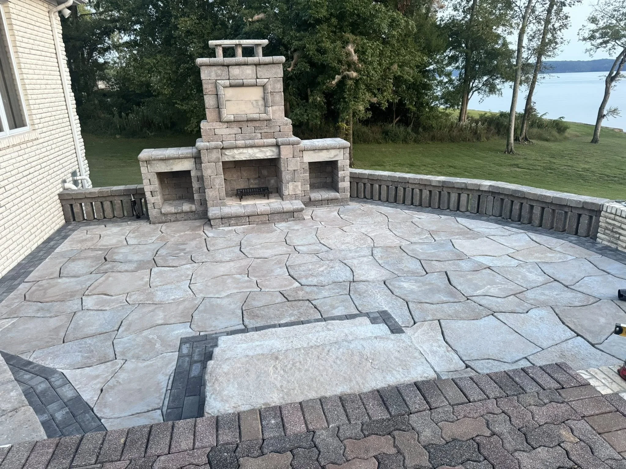 Backyard patio with stone flooring, outdoor fireplace, and a brick wall, overlooking a grassy area with trees and a body of water in the distance.