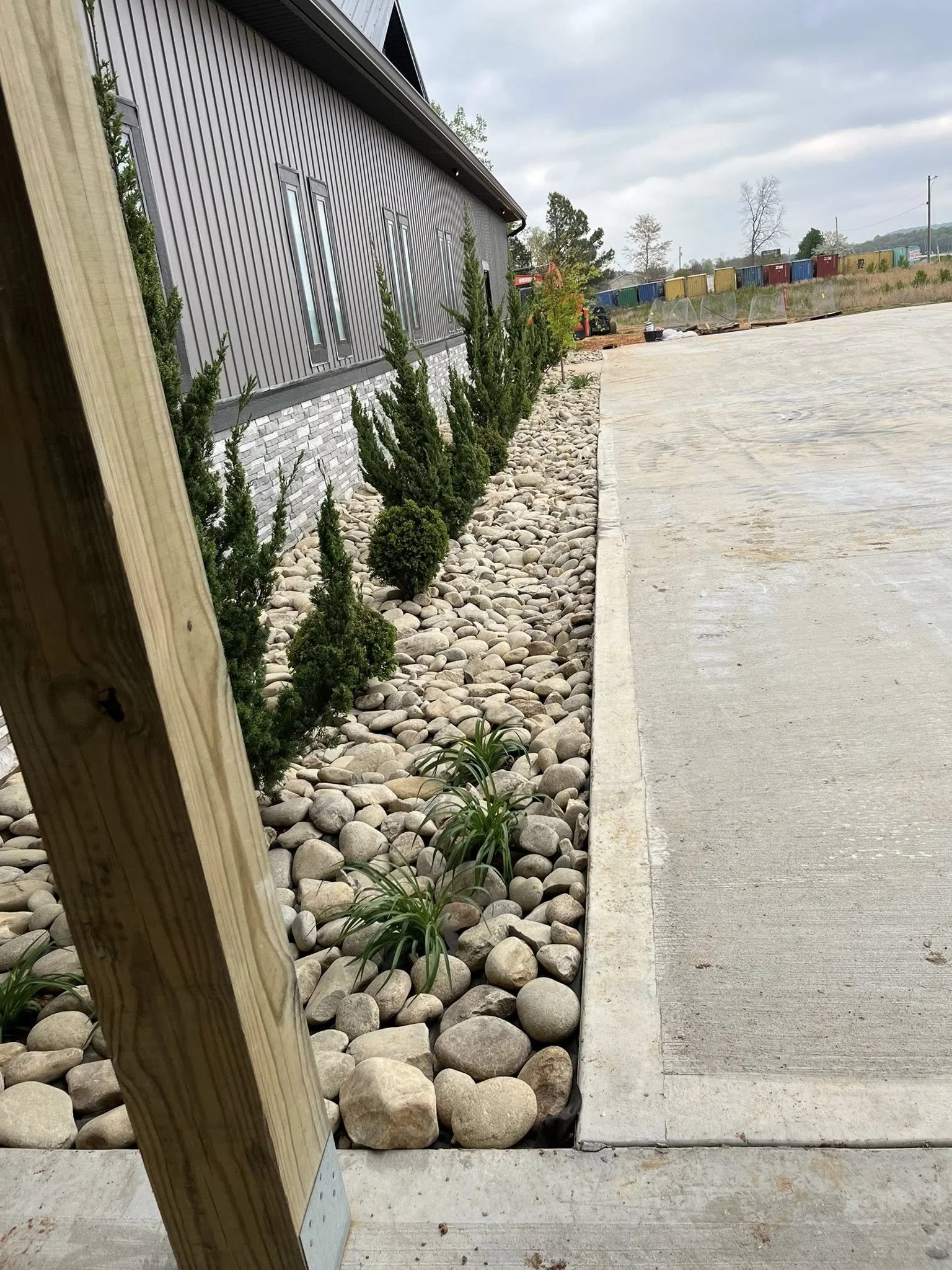 Newly landscaped area with decorative rocks and small green shrubs next to a modern gray building with metal siding and vertical windows, adjacent to a concrete sidewalk, under cloudy sky.