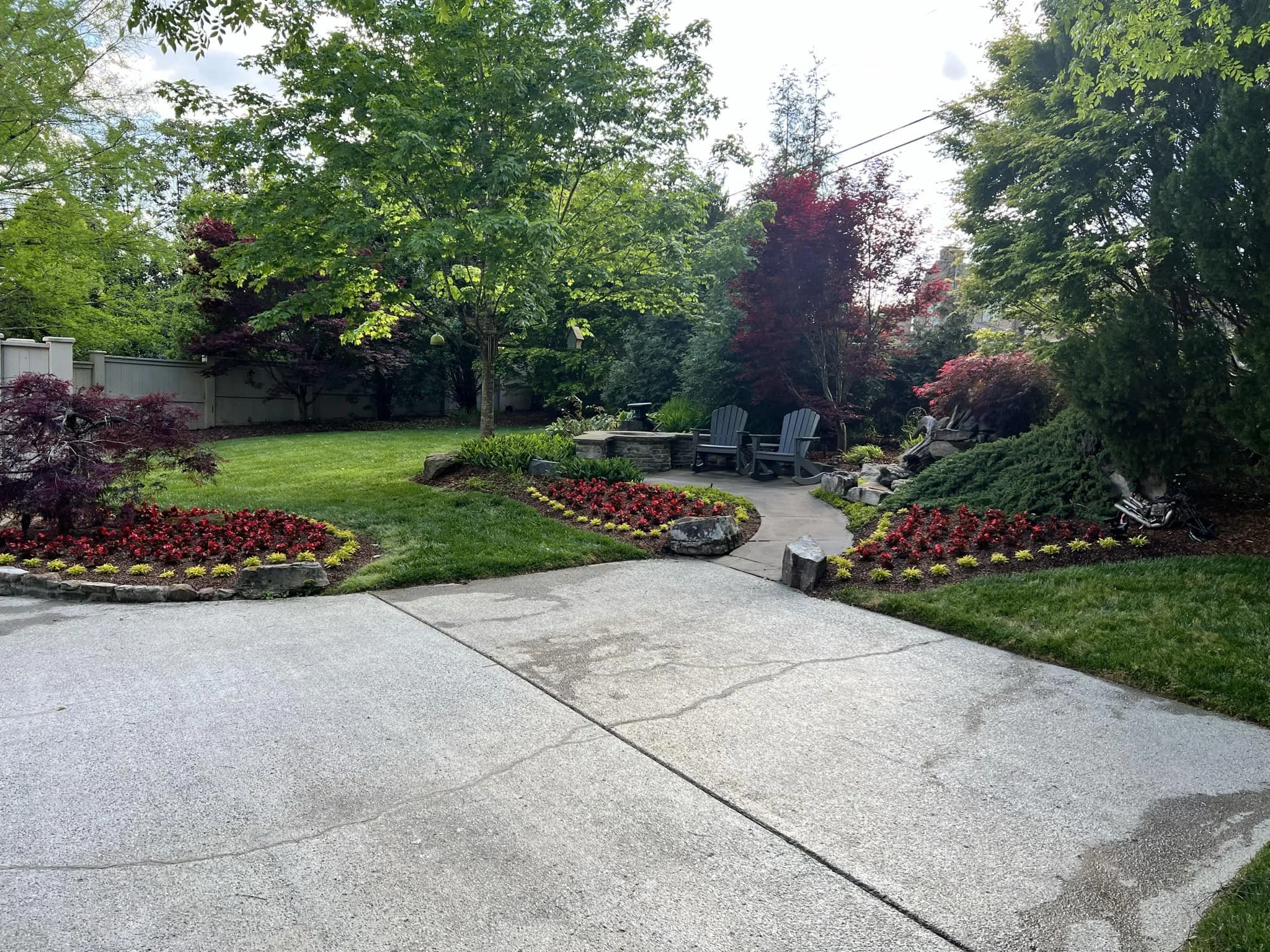 A landscaped backyard with a concrete patio, a grassy area, flower beds with red and yellow flowers, green shrubs, trees with red and green foliage, a stone seating area with two Adirondack chairs, rocks, and a fence in the background.