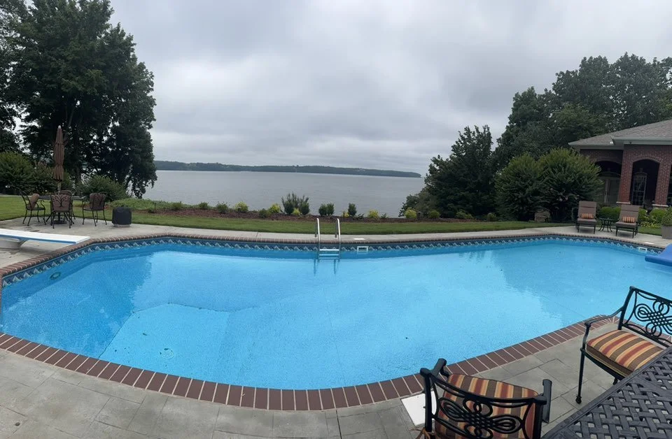 Empty swimming pool overlooking a lake with trees and cloudy sky in the background.