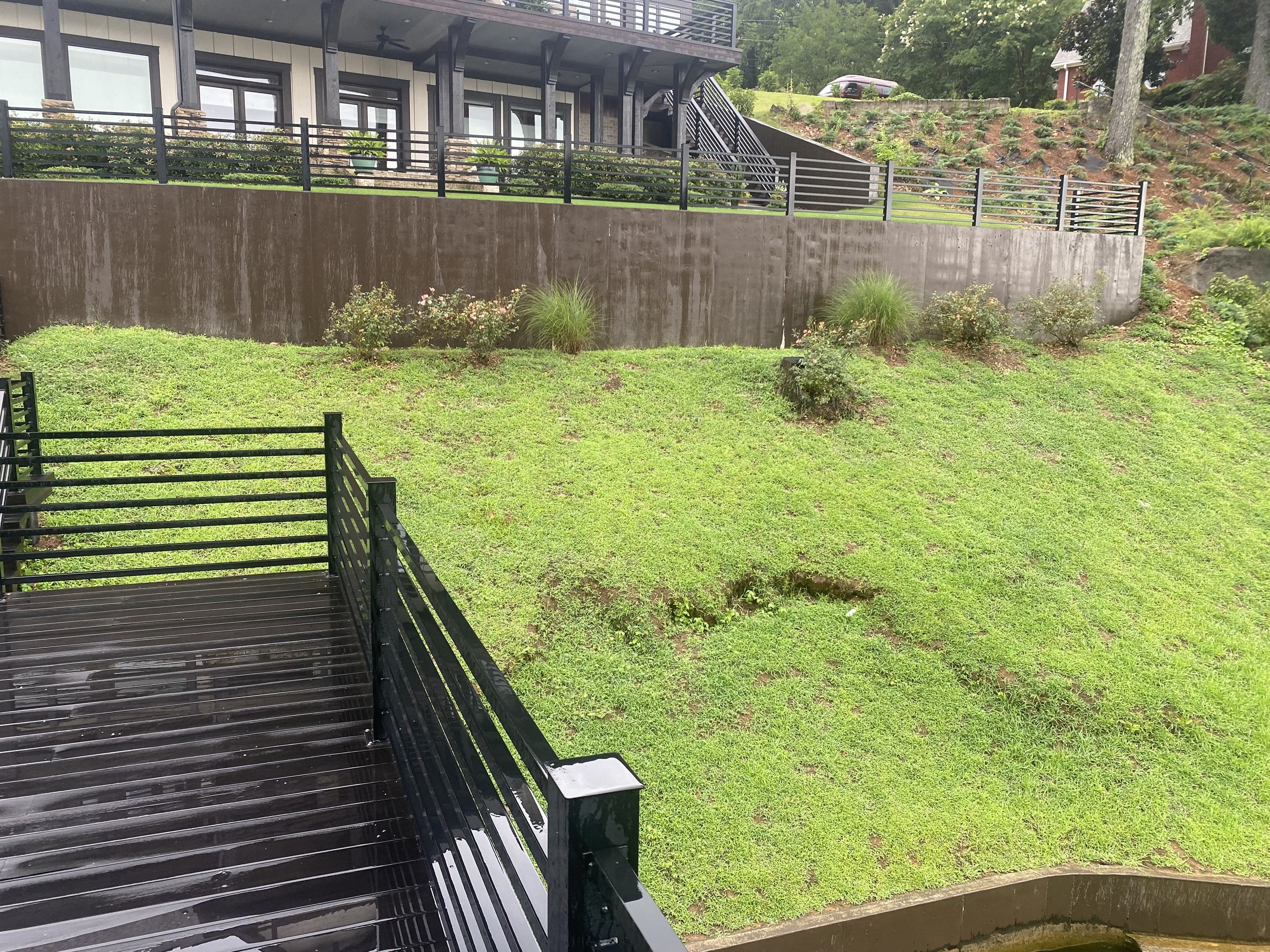Wet black metal balcony railing and floor overlooking a grassy hillside with small bushes and a concrete retaining wall, with a house and trees in the background on a rainy day.