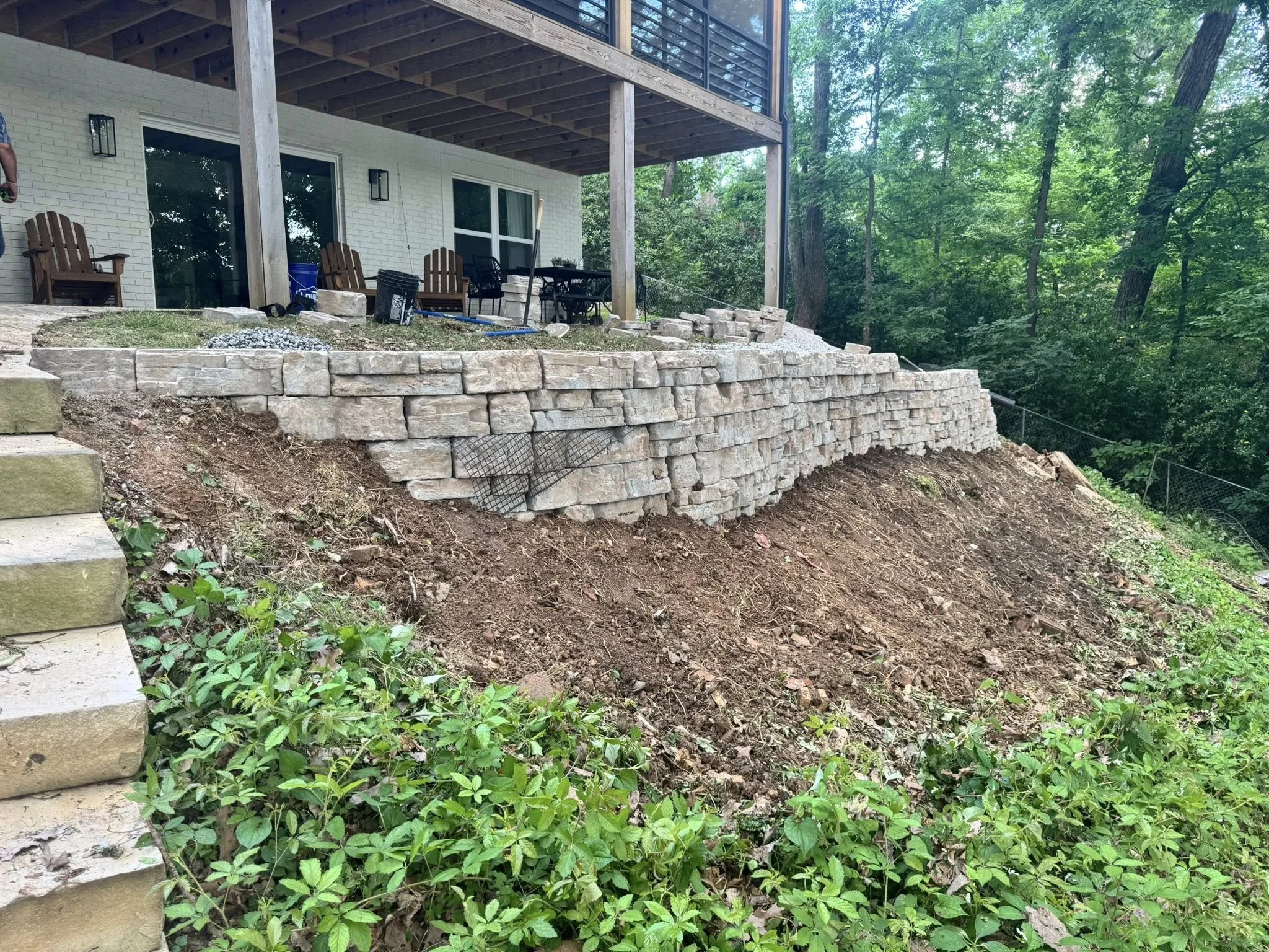 A back porch of a house with a stone retaining wall under construction on a hill, surrounded by trees and greenery.