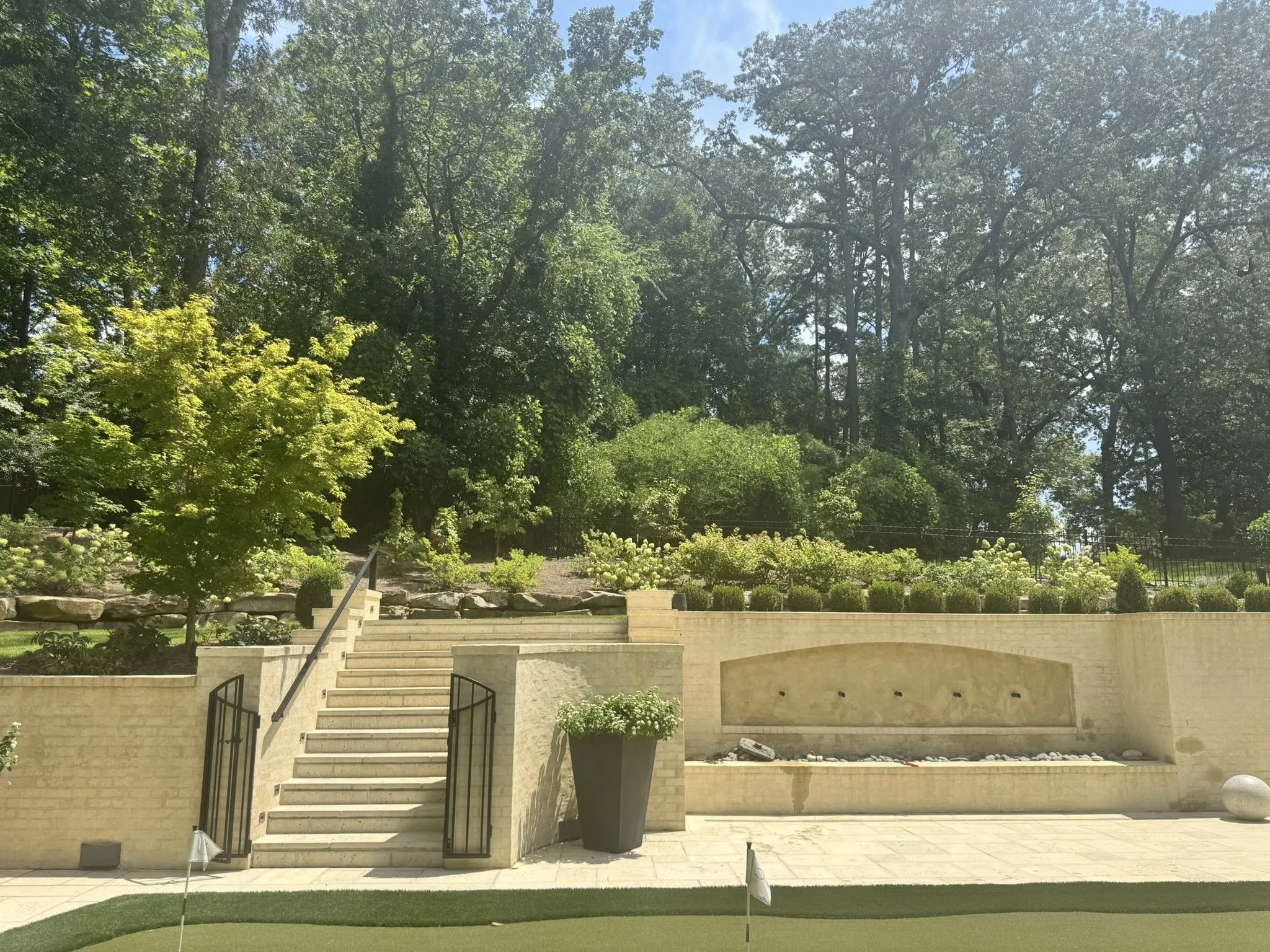 A backyard with stairs, a water feature, a railing, and lush green trees and plants under a clear blue sky.