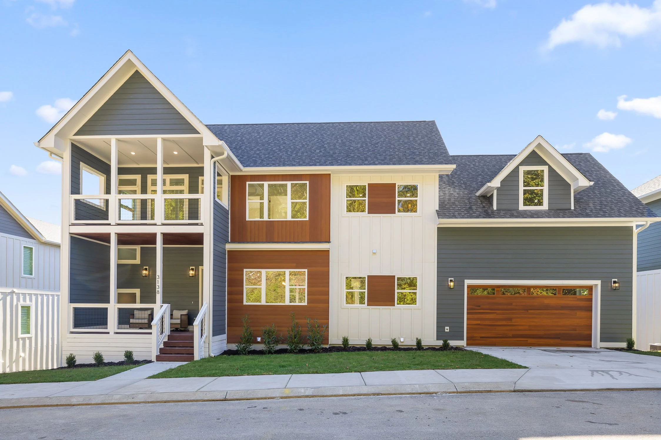Front view of a modern multi-story house with navy blue, white, and wood paneling exterior, a two-car garage with a wooden door, and a small front porch with seating. Clear sky with a few clouds in the background.
