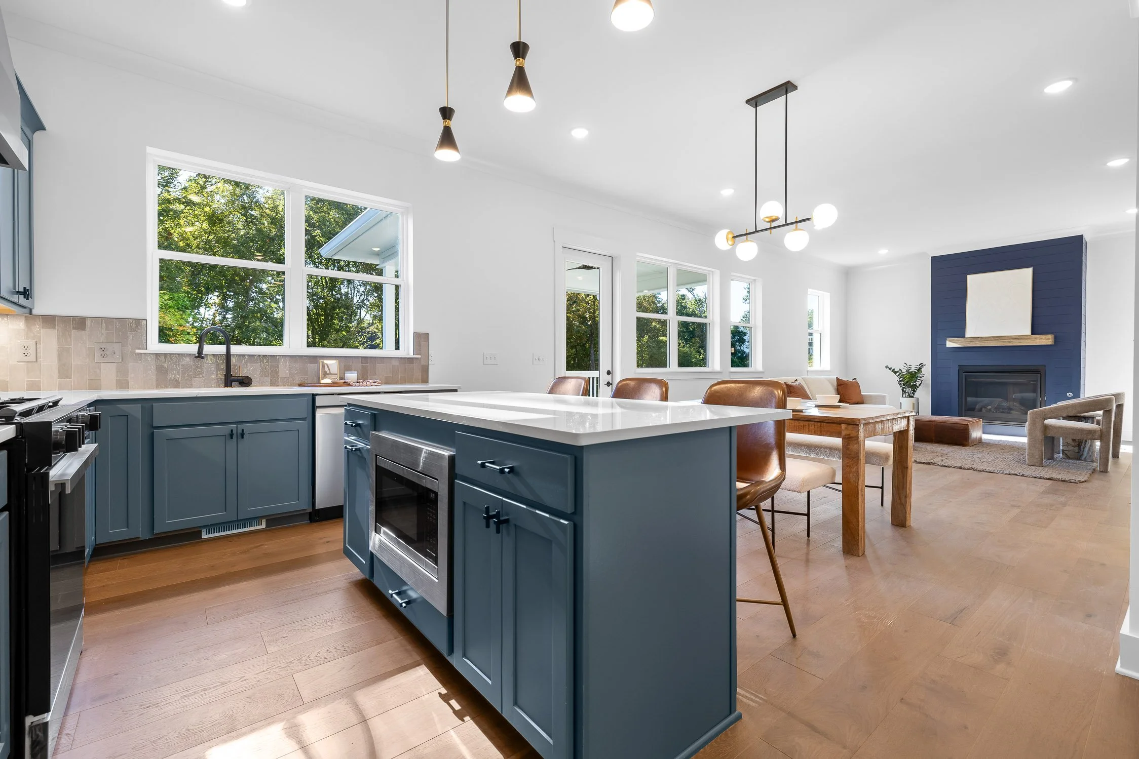 Open-concept kitchen and living room with blue cabinets, wooden dining table, leather chairs, and a blue accent wall with a fireplace and mirror.