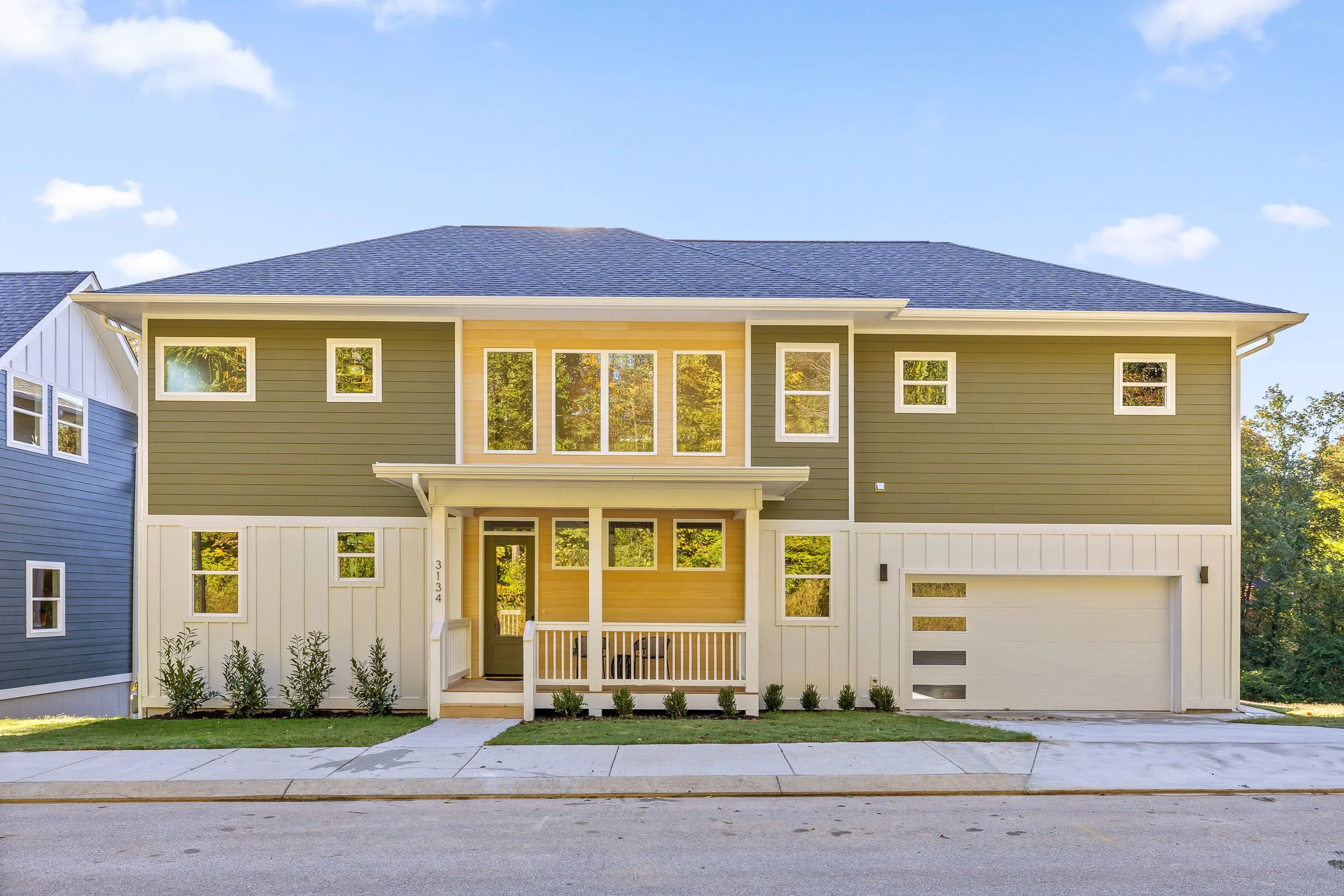 Modern two-story house with green and beige siding, front porch, and attached garage, surrounded by a small lawn and shrubs, under a blue sky.